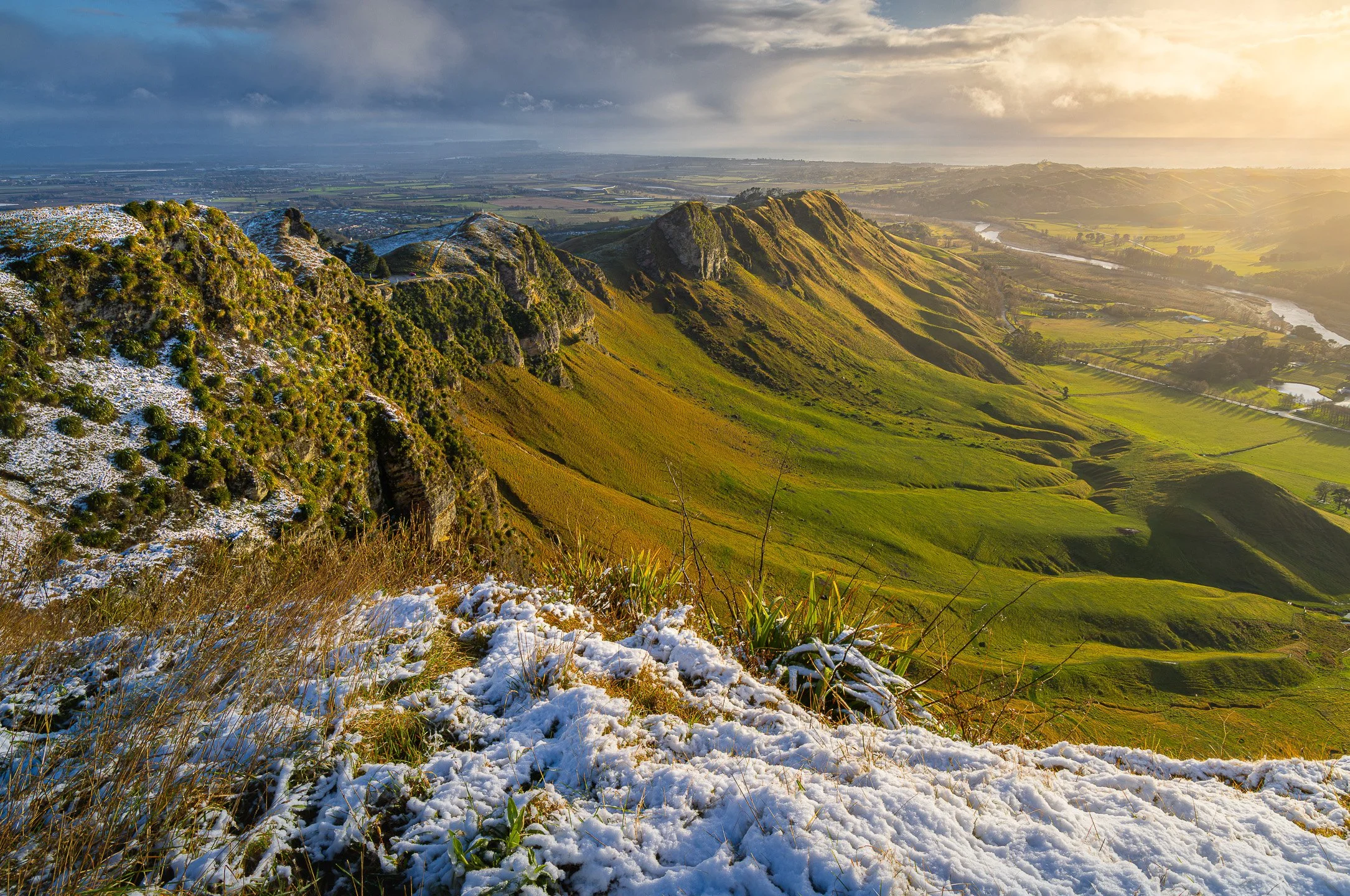 Rocky ridge on Te Mata Peak dusted with snow, with green farmland stretching out below in morning light.