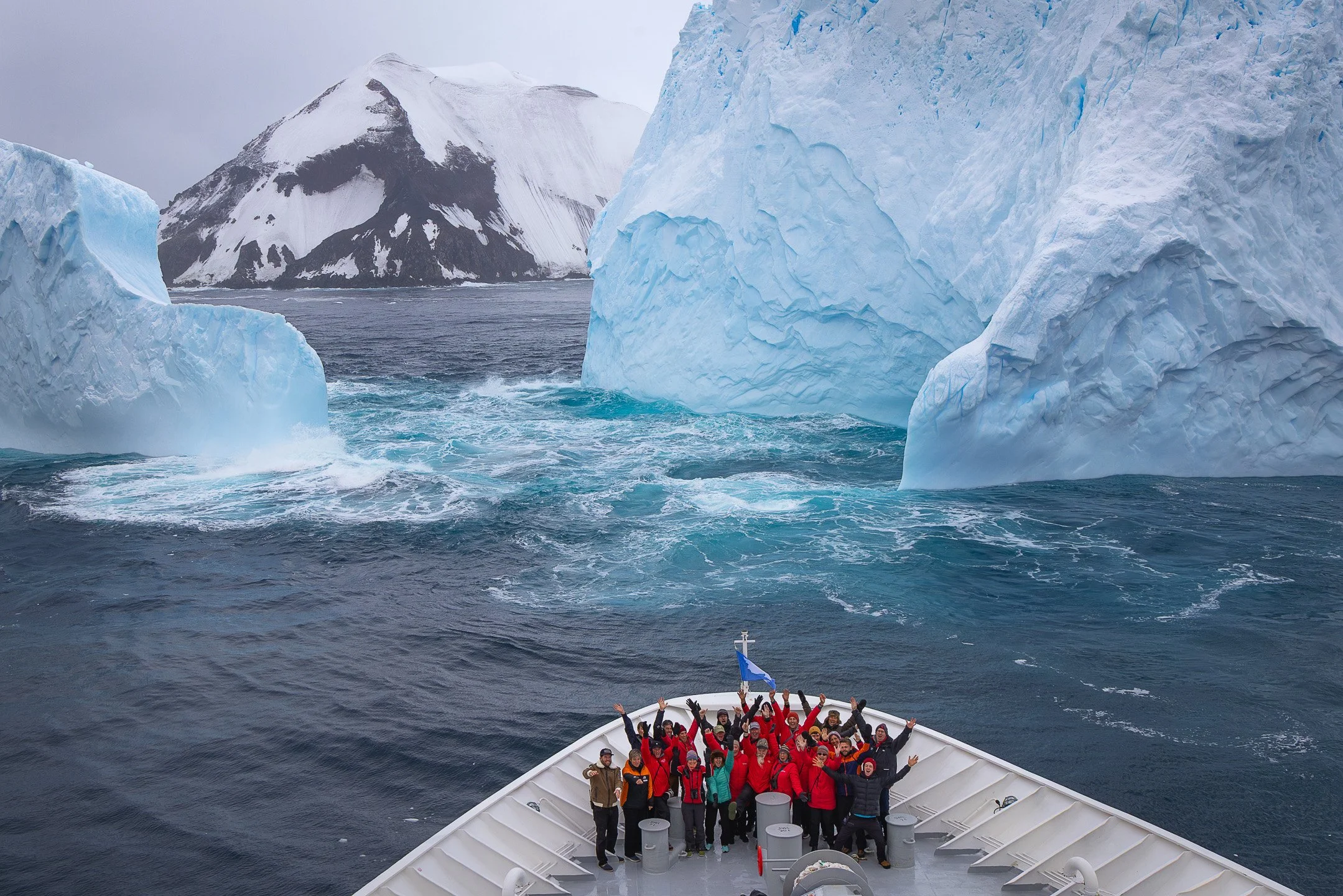 The expedition team gathered on the bow of the ship at the Balleny Islands, Antarctica, surrounded by towering icebergs and volcanic peaks, arms raised in celebration.