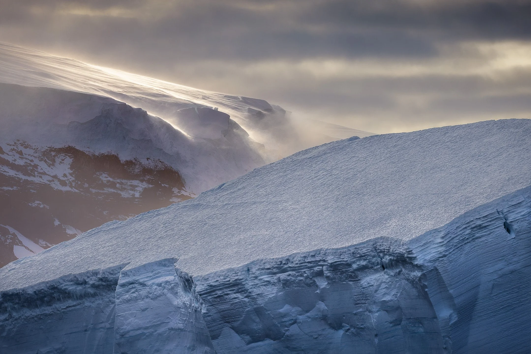 Fierce light pulls back toward the horizon over a glacial slope at the Balleny Islands, Antarctica, leaving a warm diffuse glow while an iceberg catches the last of it in the foreground.