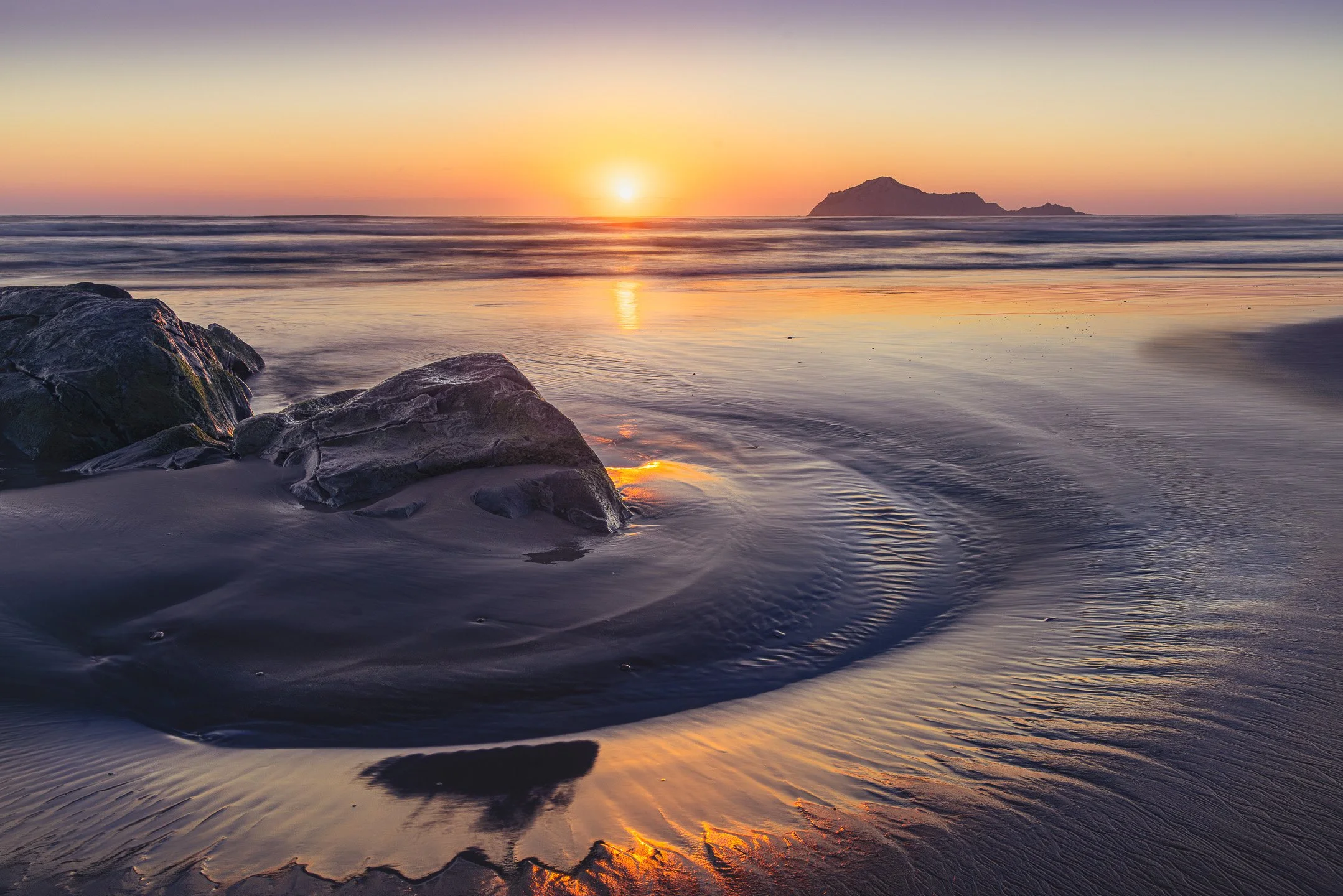 Golden sunrise light reflects off tide-carved wet sand at Waimārama Beach, dark weathered rocks in the foreground and the hazy silhouette of Bare Island on the horizon, Hawke's Bay, New Zealand.