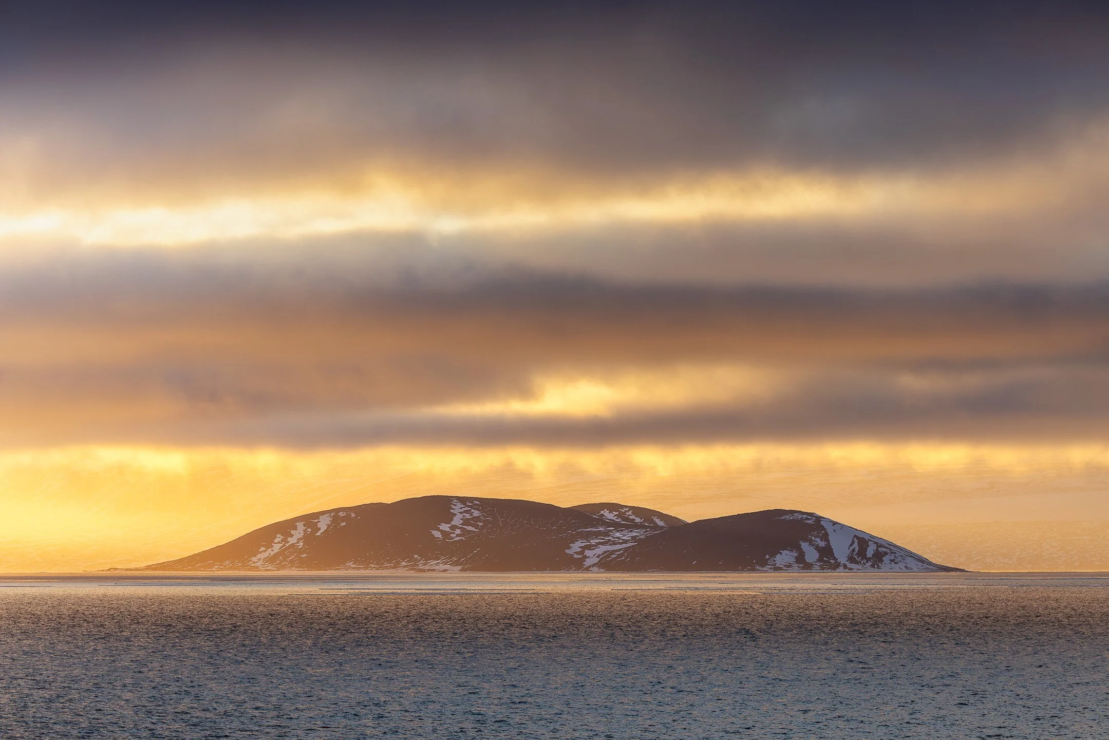 A dark, snow-dusted island sits beneath a dramatic sky of layered, golden-lit clouds reflecting on the Antarctic sea.