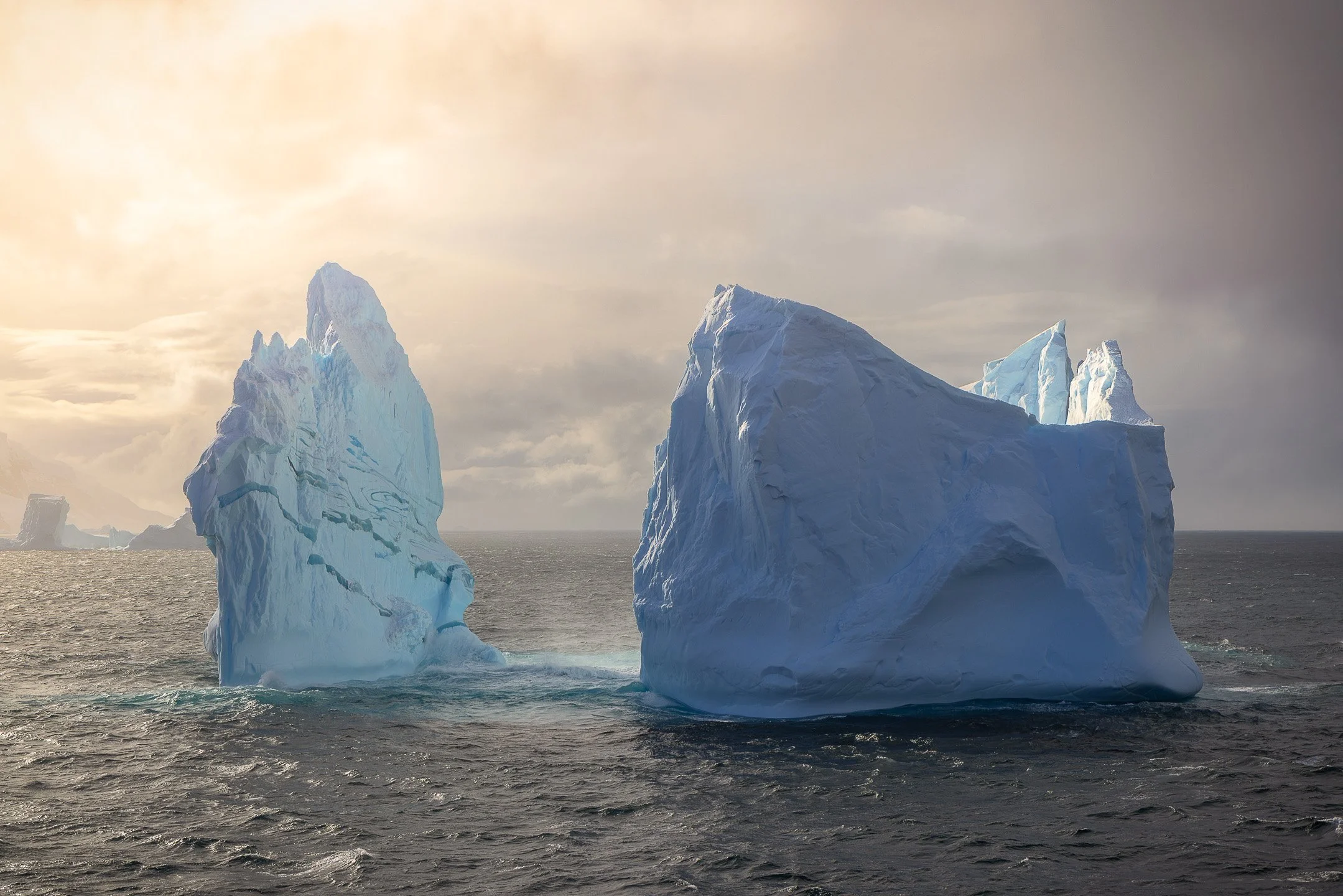 Two icebergs in the Southern Ocean near the Balleny Islands bathed in warm golden light, the same forms photographed earlier in grey overcast conditions now completely transformed by the breaking sun.