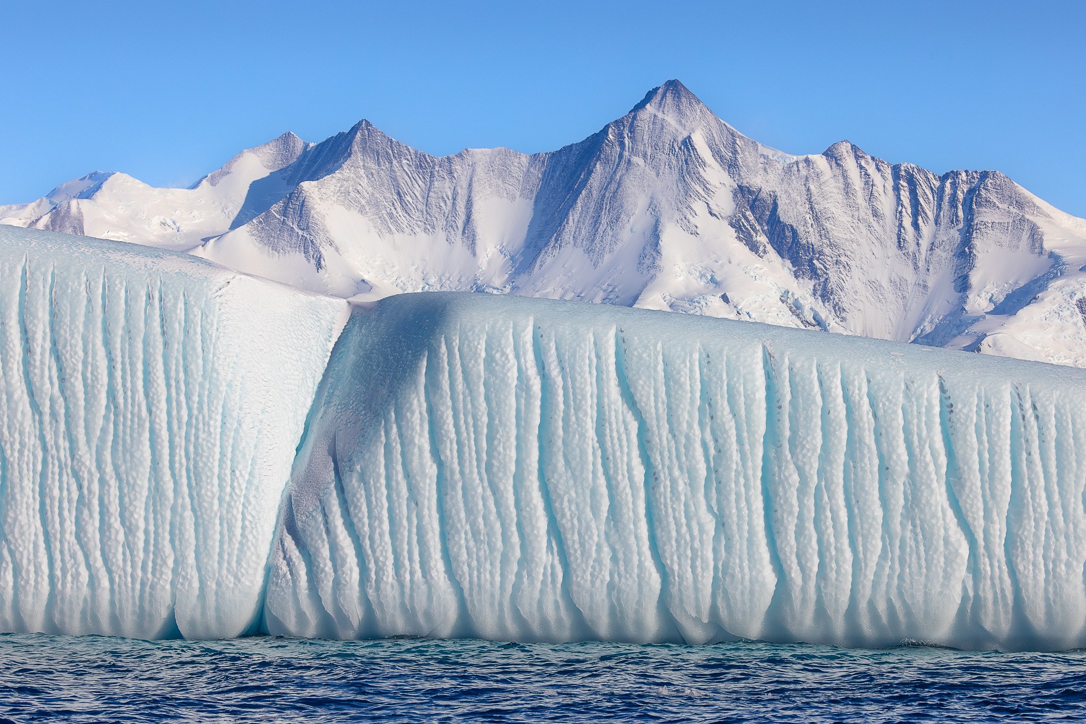A massive, fluted iceberg wall with vertical ridges sits in front of jagged, snow-covered mountains under a clear blue sky.