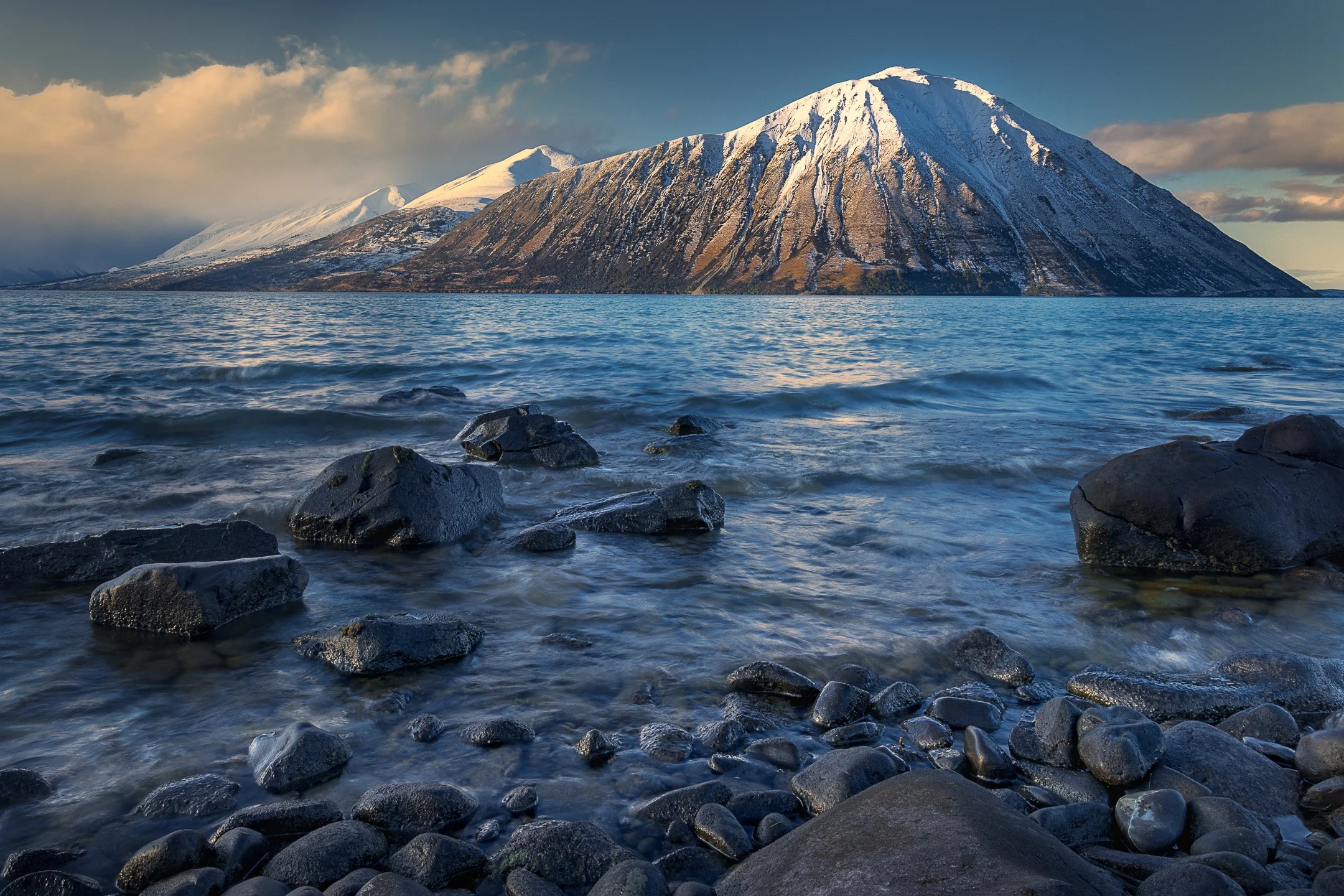 Ben Ohau dominates the southern end of Lake Ōhau, a grey foreground rock echoing the shape of the snow-covered peak behind it, the lake surface calm in the cool morning light, Canterbury, New Zealand.
