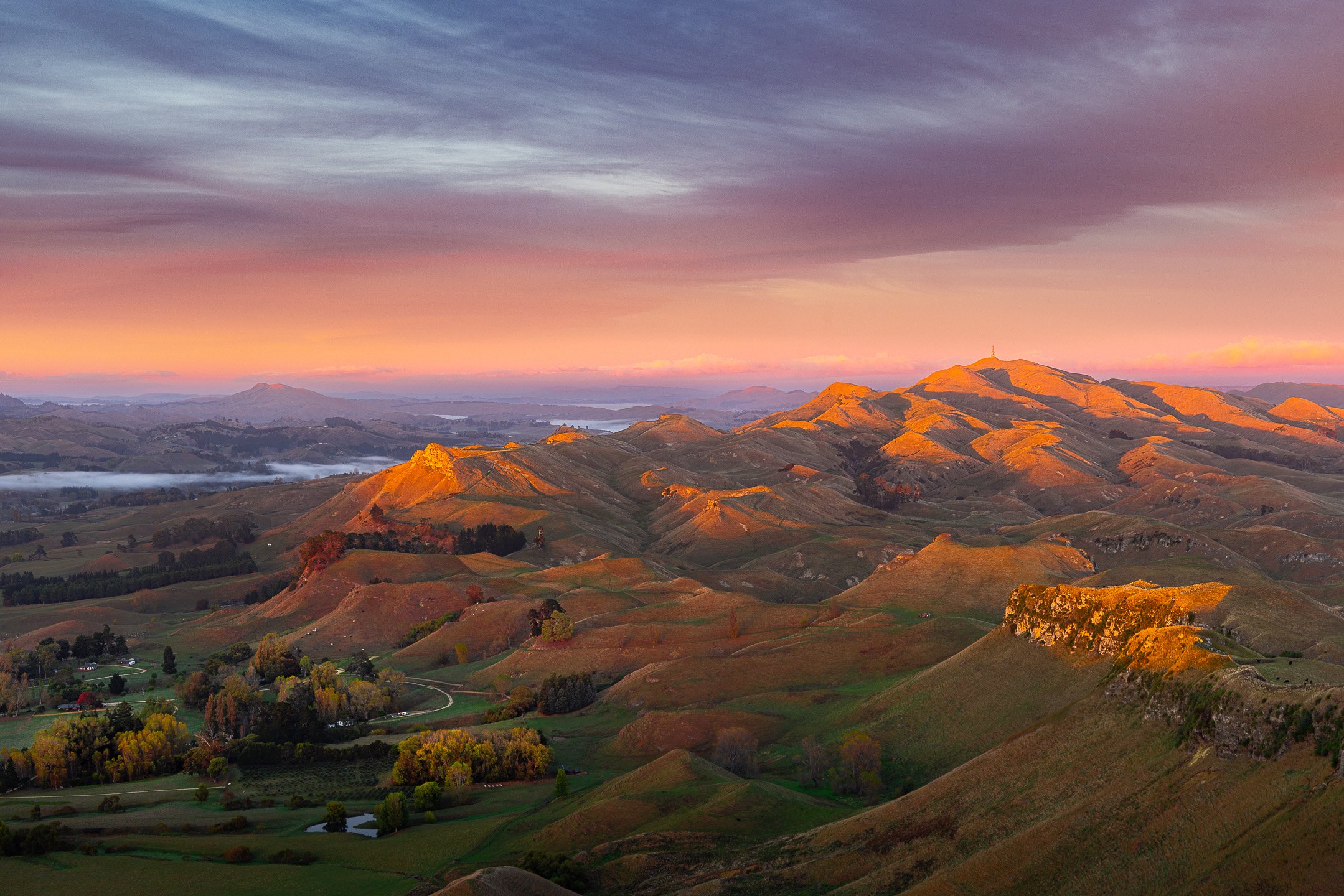 Golden dawn light hitting Mount Erin and rolling hill country in Hawke's Bay, New Zealand, with fog pooled in a distant valley beneath a pink and purple sky.