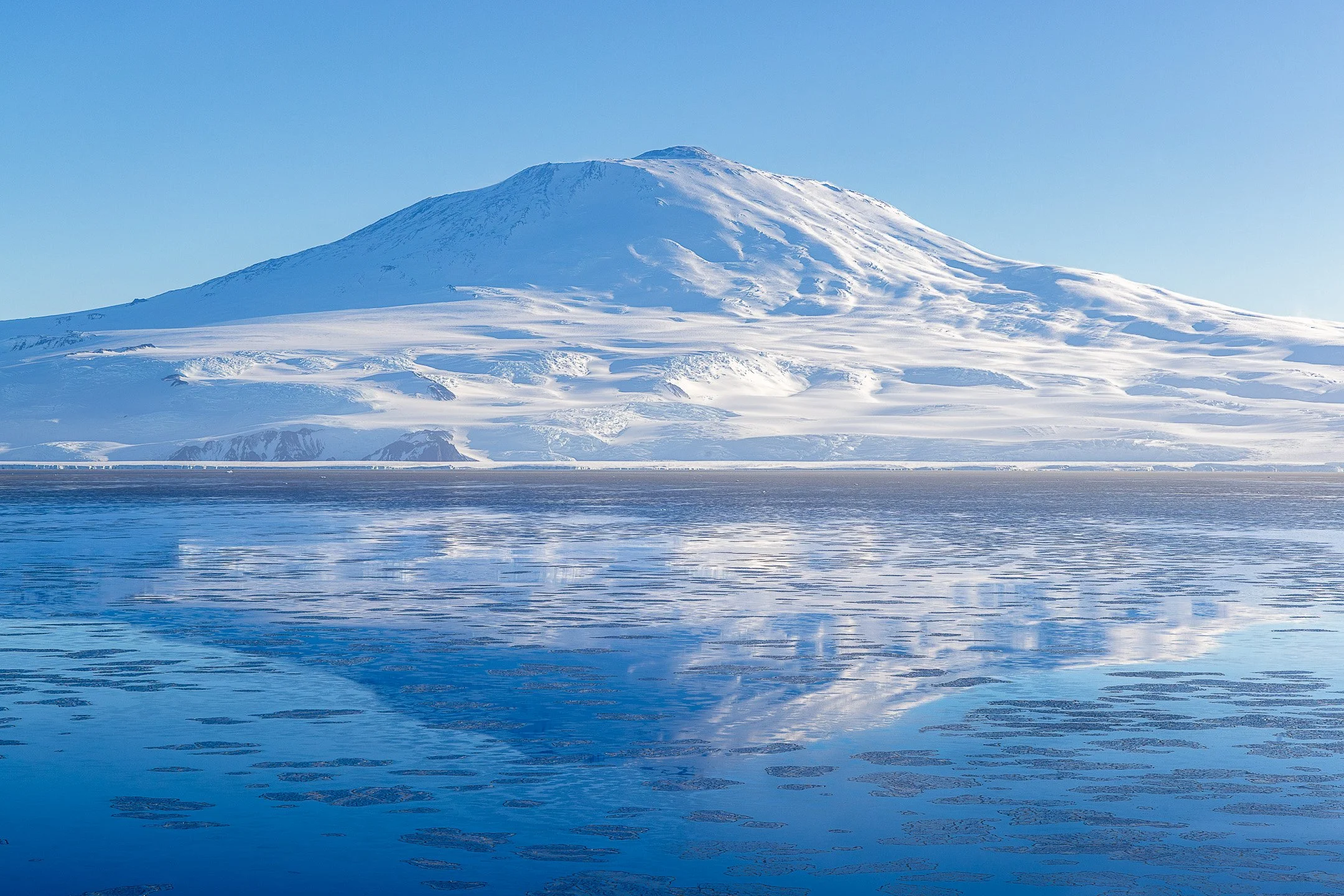 Mount Erebus, Antarctica's active volcano, rises above McMurdo Sound in pale blue winter light, its snow-covered flanks reflected in the near-still surface of the Ross Sea, broken only by scattered fragments of pancake ice.