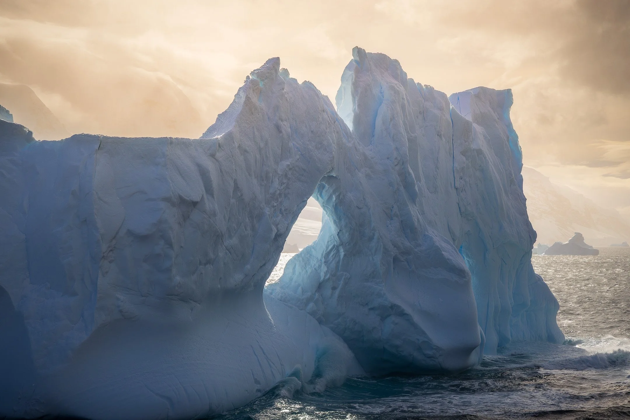 Close view of a monumental ice arch near the Balleny Islands, Antarctica, with one face bright in golden light and one in shadow, the opening between them framing the Southern Ocean beyond.
