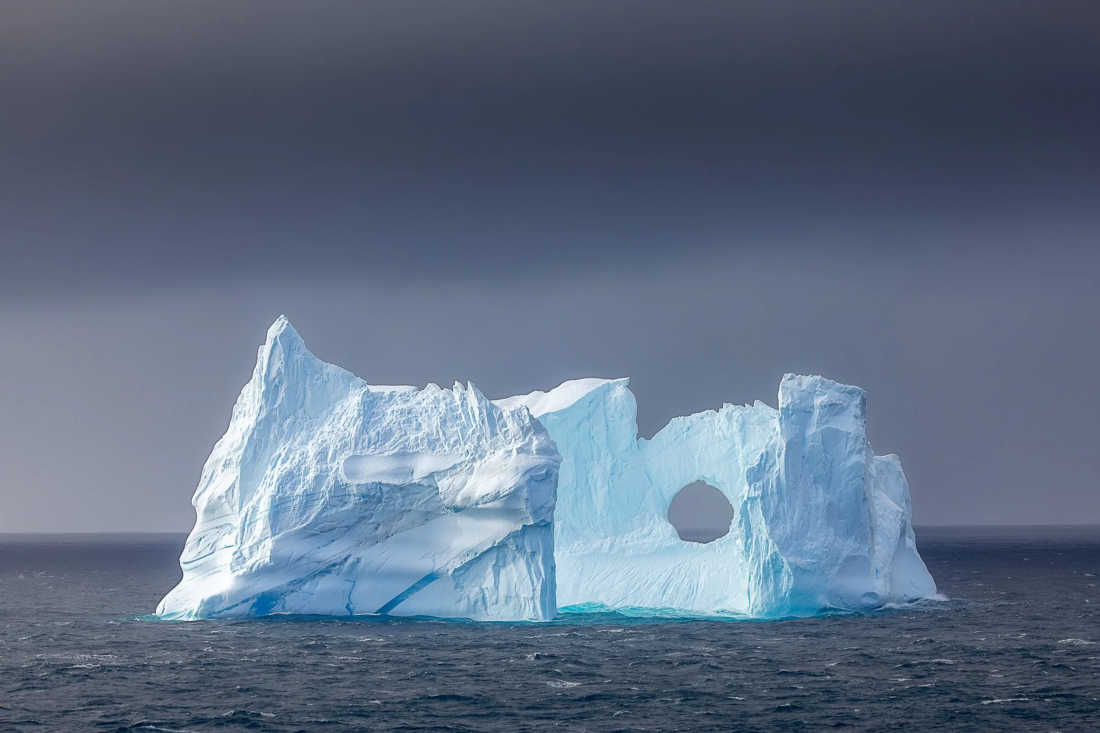A perfect circular opening worn through the body of a large iceberg in the Southern Ocean near the Balleny Islands, Antarctica — a natural arch created by time and water.