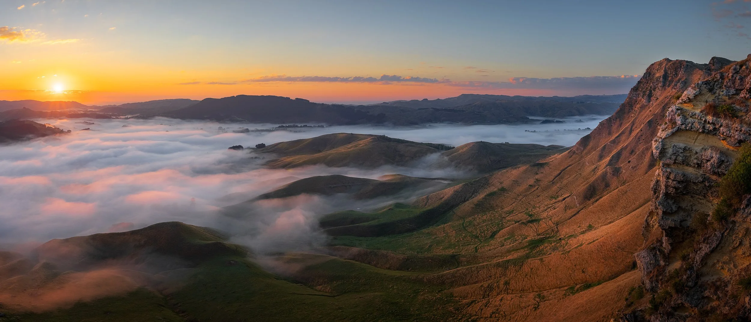 Panoramic sunrise view over the Tukituki Valley filled with fog, with rocky cliffs and rolling hills rising above the cloud, Hawke's Bay, New Zealand.