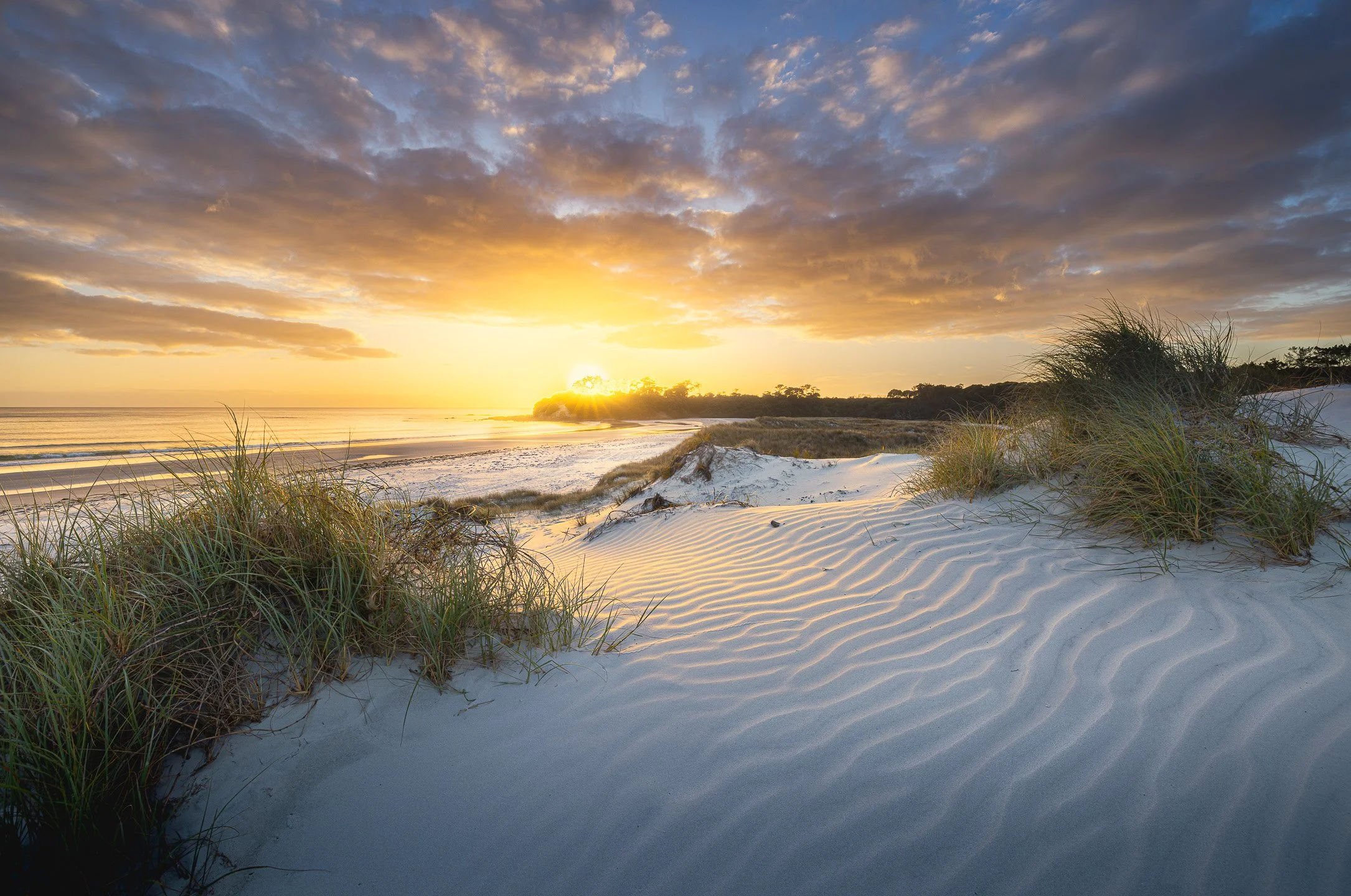 Pristine white sand dunes at Rarawa Beach catch the warm golden light of dawn, rippling sand patterns illuminated beneath a sky shifting from deep orange to pale blue, Northland, New Zealand.