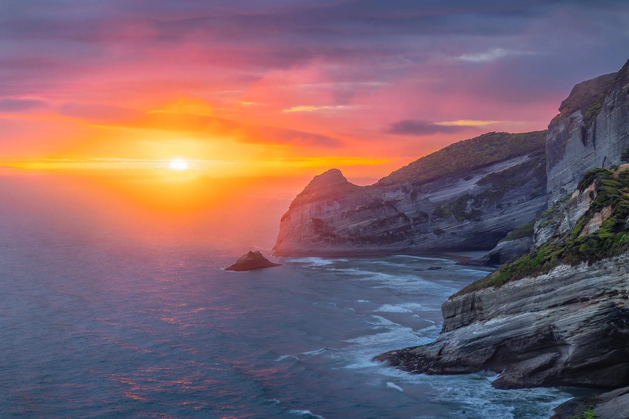 Red sun glowing through sea mist over cliffs near Cape Farewell, Puponga, Tasman, New Zealand