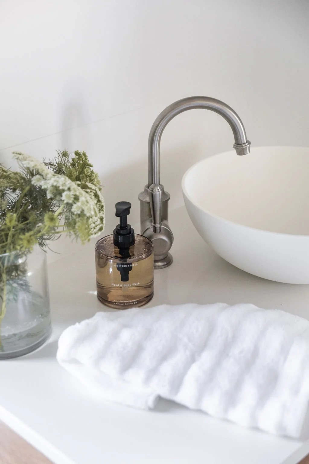 A white bathroom sink with a silver faucet, a brown hand and body wash bottle, a white towel, and a glass vase with green and white flowers.