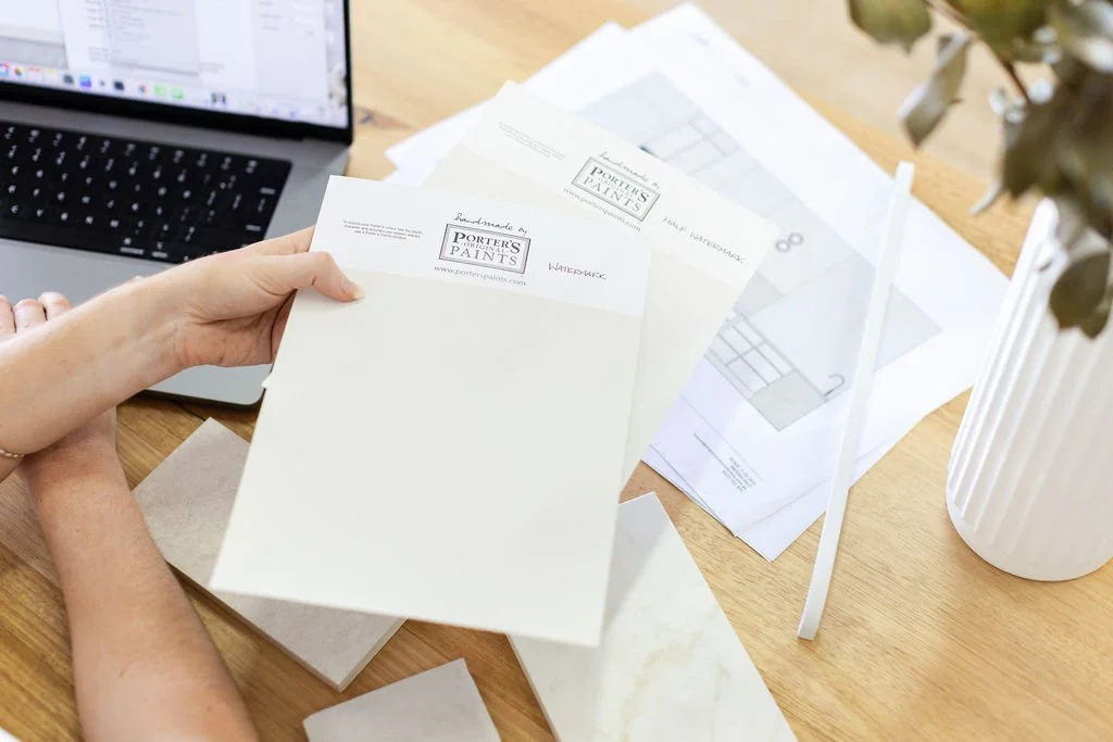Person holding custom watercolor paint label paper with a laptop and architectural plans on a wooden desk.