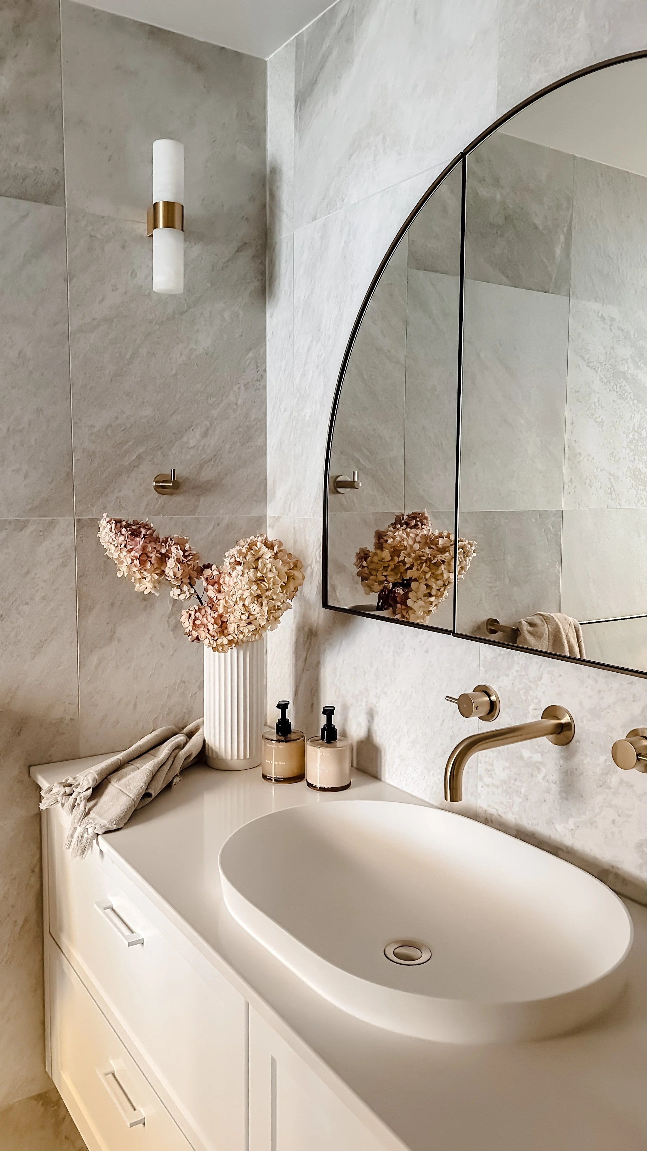 Modern bathroom vanity with a white countertop, oval sink, gold fixtures, large mirror, beige floral arrangement in a white vase, soap dispensers, and neutral-toned wall tiles.