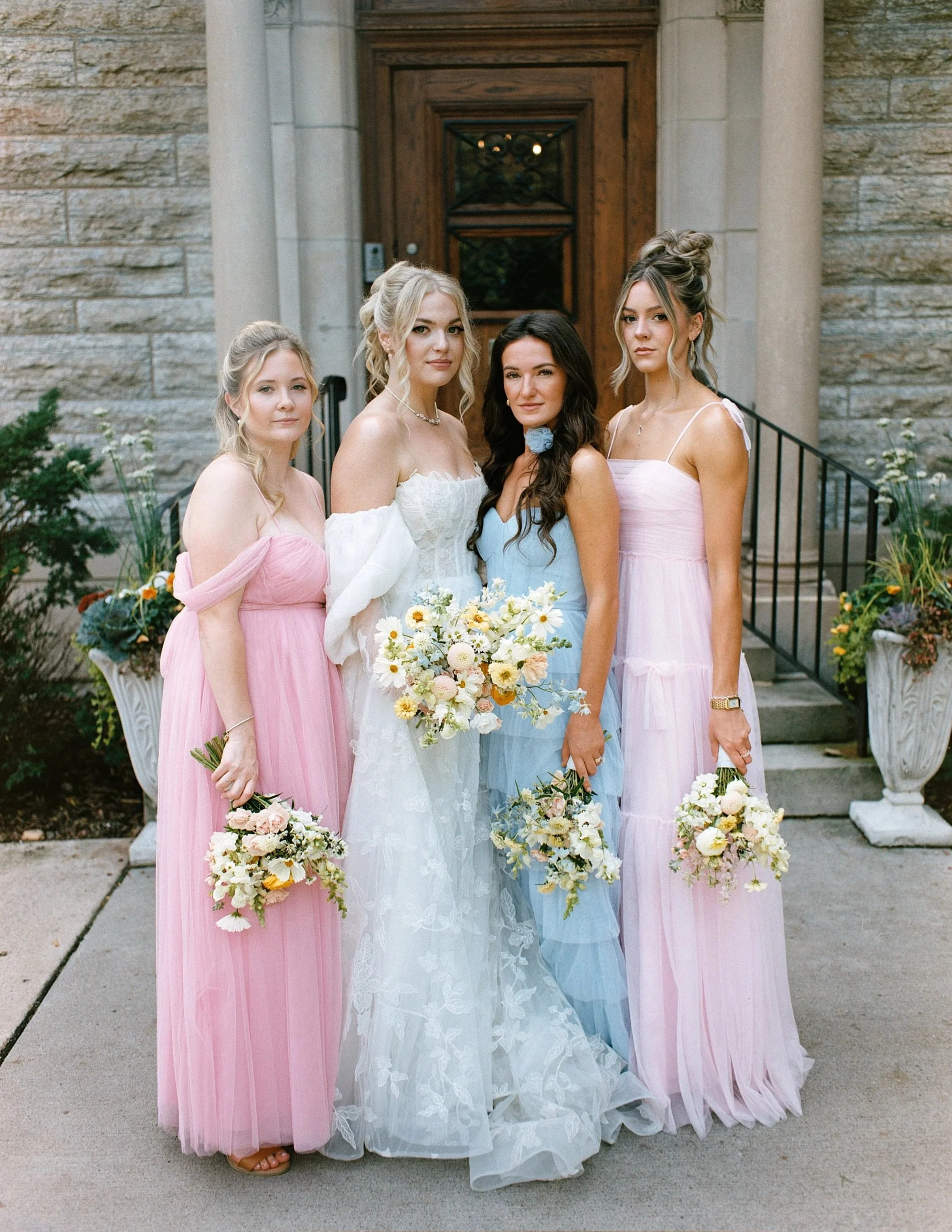 A bride in a white wedding dress surrounded by four women in pastel-colored dresses holding bouquets, standing in front of a stone building with potted plants.