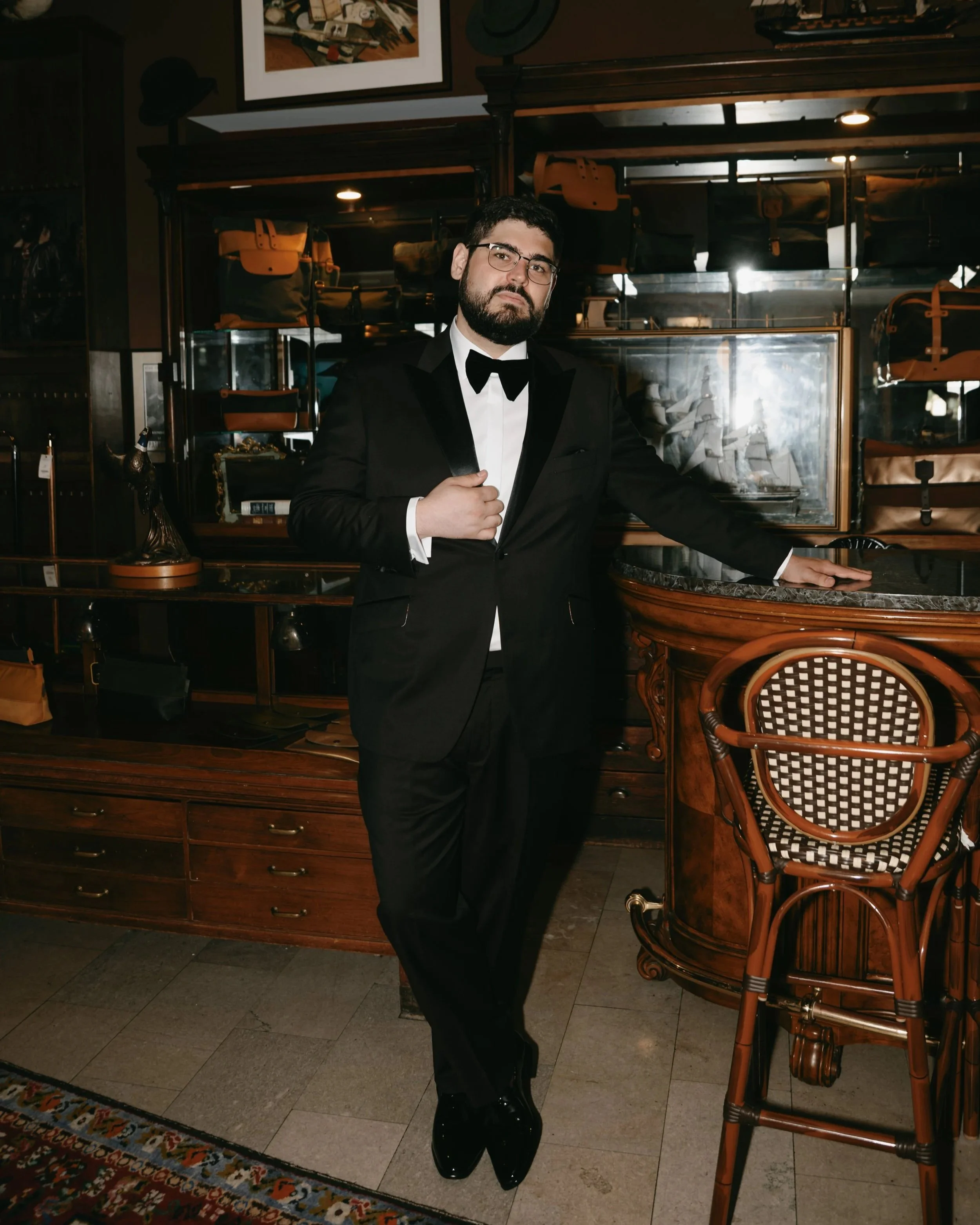 A man dressed in a black tuxedo with a bow tie, glasses, and a beard poses indoors next to a wooden bar and wicker chair, with shelves of bags and artwork in the background.