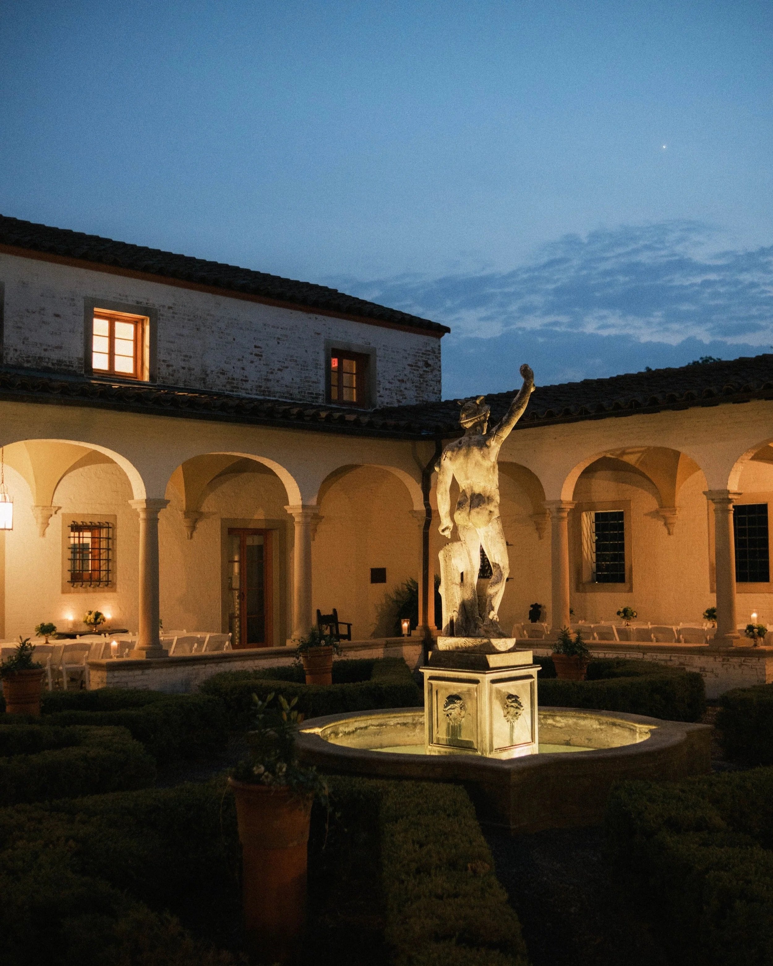A courtyard with a fountain and statue of a man, lit by warm lights at dusk, surrounded by potted plants and arches.