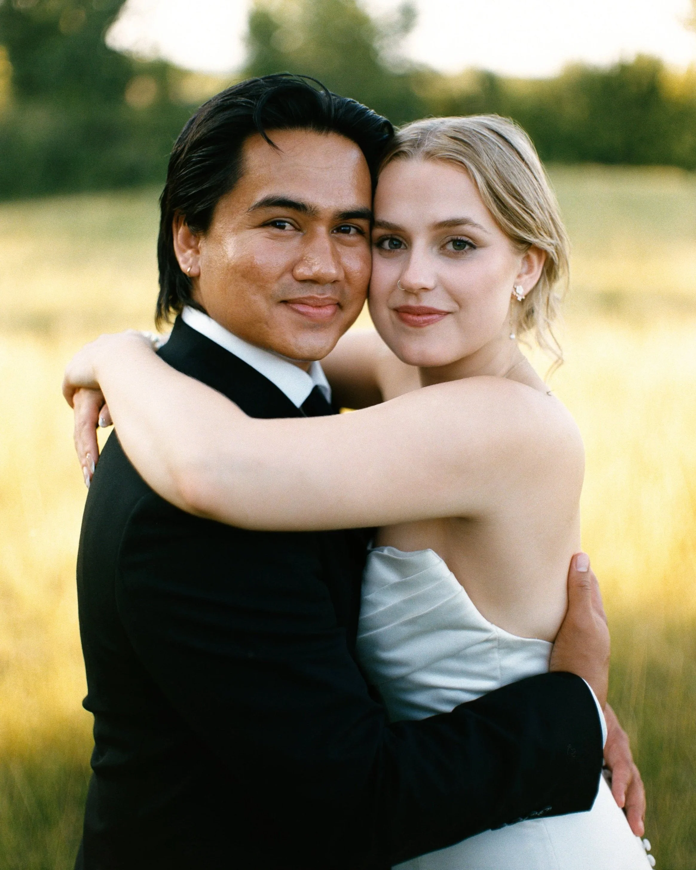 A couple embracing outdoors during daytime, with trees and a green field in the background.