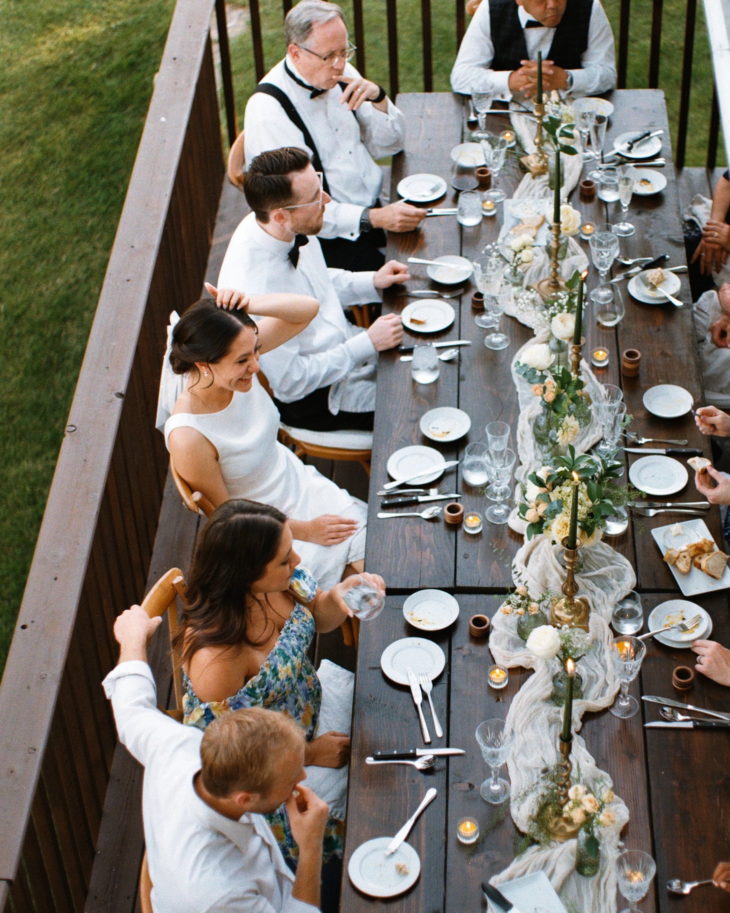 People gathered at a long outdoor banquet table decorated with flowers and candles during a formal event, possibly a wedding reception.