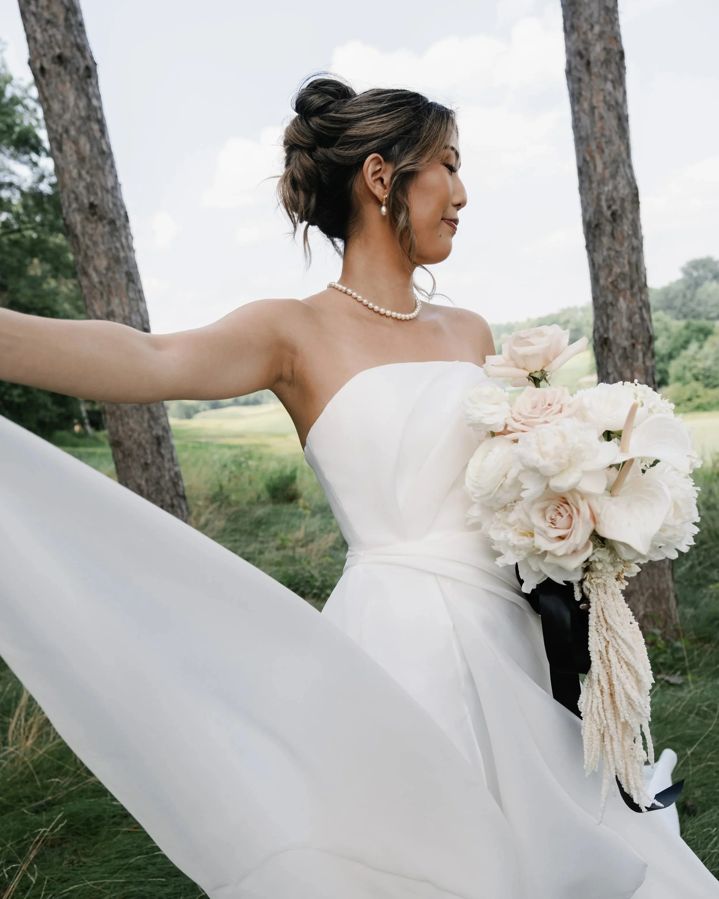 A bride in a white wedding dress holding a bouquet of flowers outdoors on a grassy area with trees in the background.