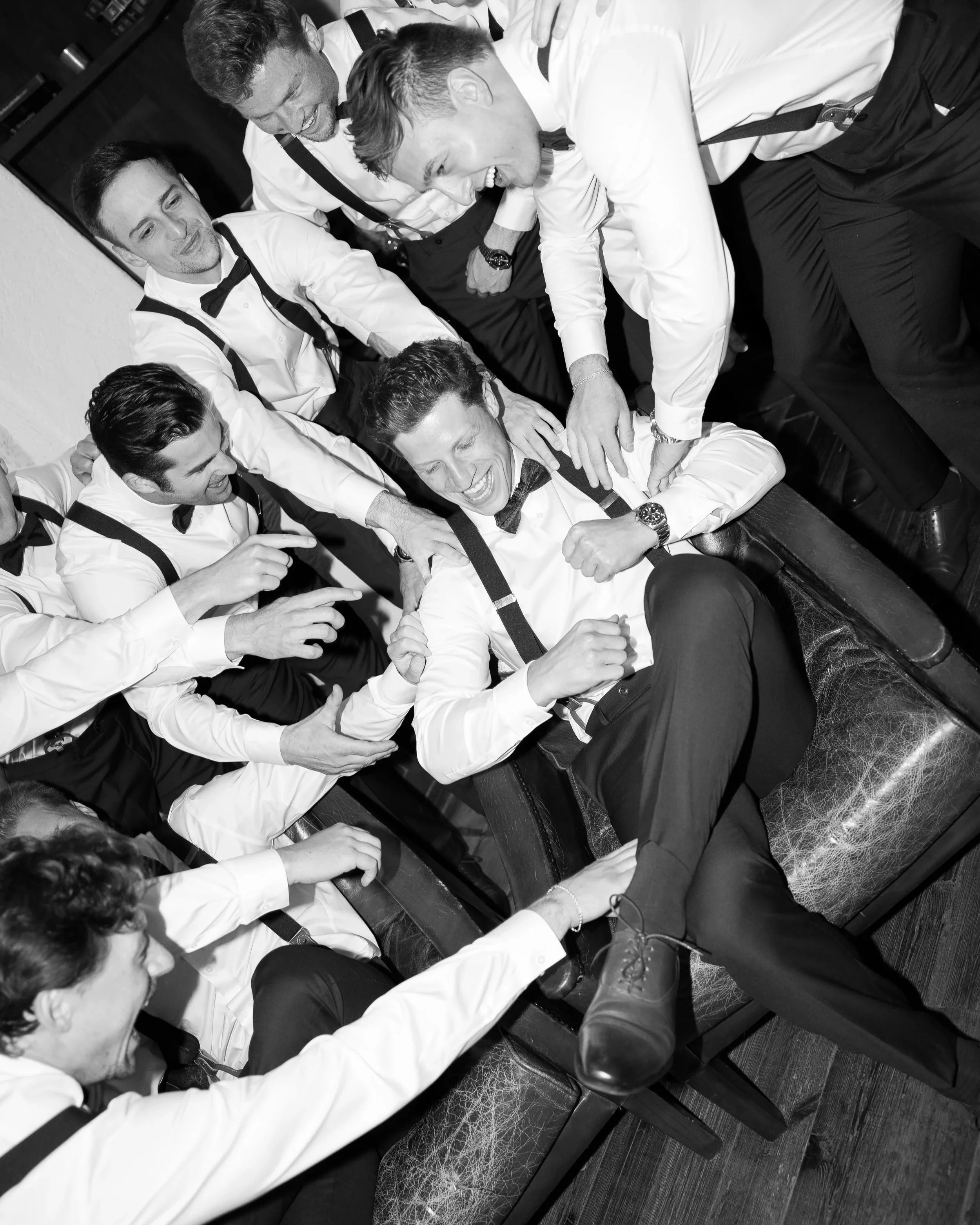 Group of men in tuxedos and suspenders playfully horsing around in a room, with some sitting and others standing, sharing laughs and engaging with each other.