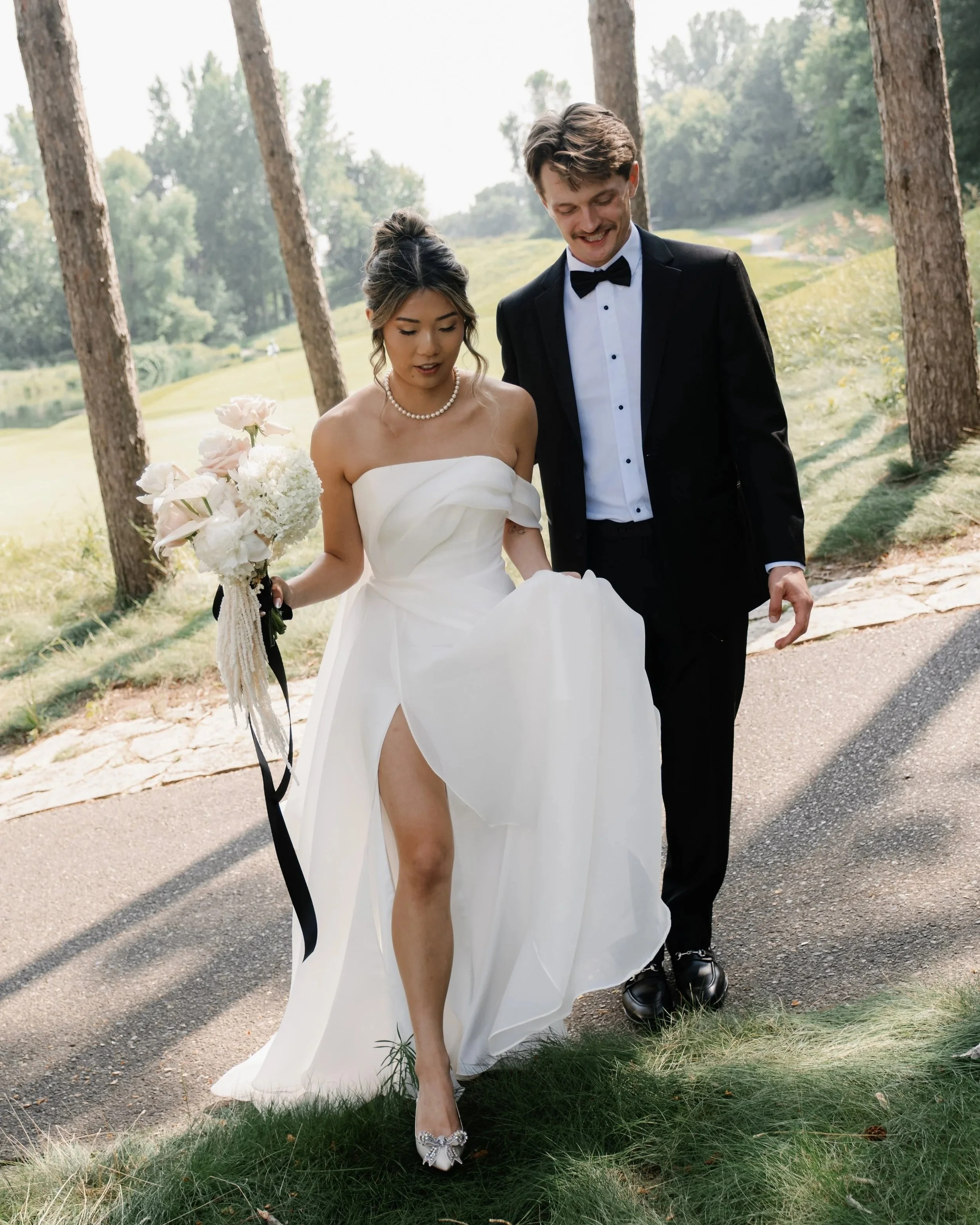 A bride wearing a white strapless wedding gown with a slit, and a groom in a black tuxedo with a bow tie, walking outdoors on a paved path with green grass and tall trees in the background.