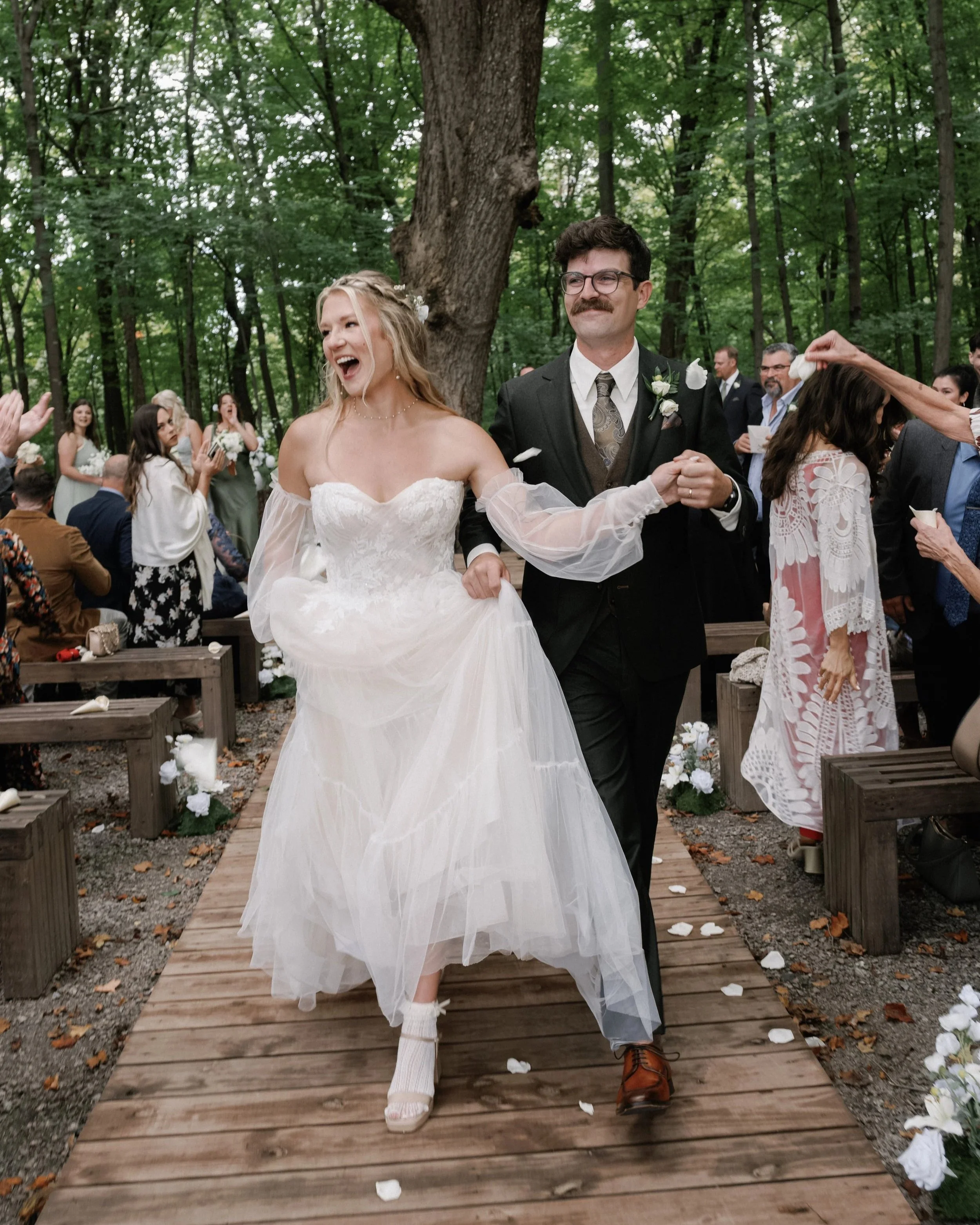 A newlywed couple walks down an outdoor aisle, surrounded by friends and family. The bride is in a strapless white wedding dress with a flowing skirt, and the groom is in a black suit with a boutonniere. They are smiling and appear joyful, with trees