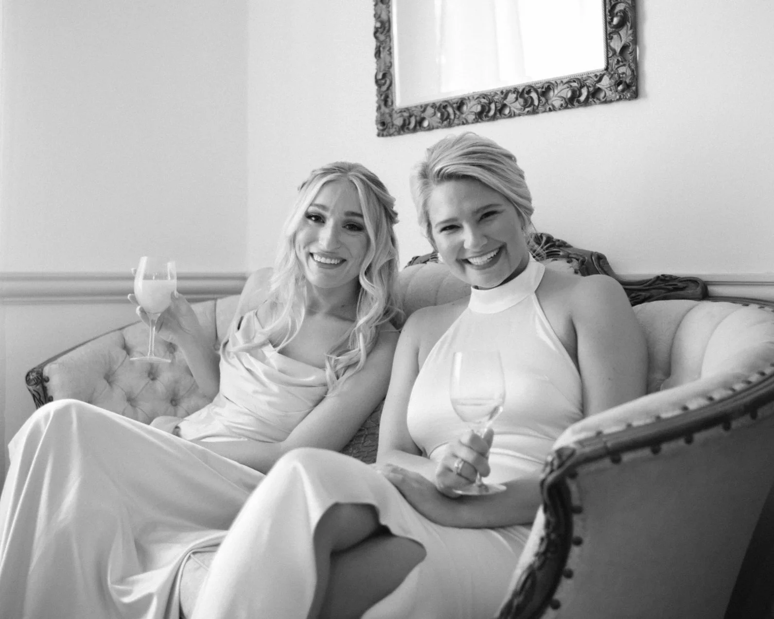 Two women in elegant dresses smiling and holding glasses on a vintage sofa in a well-lit room.