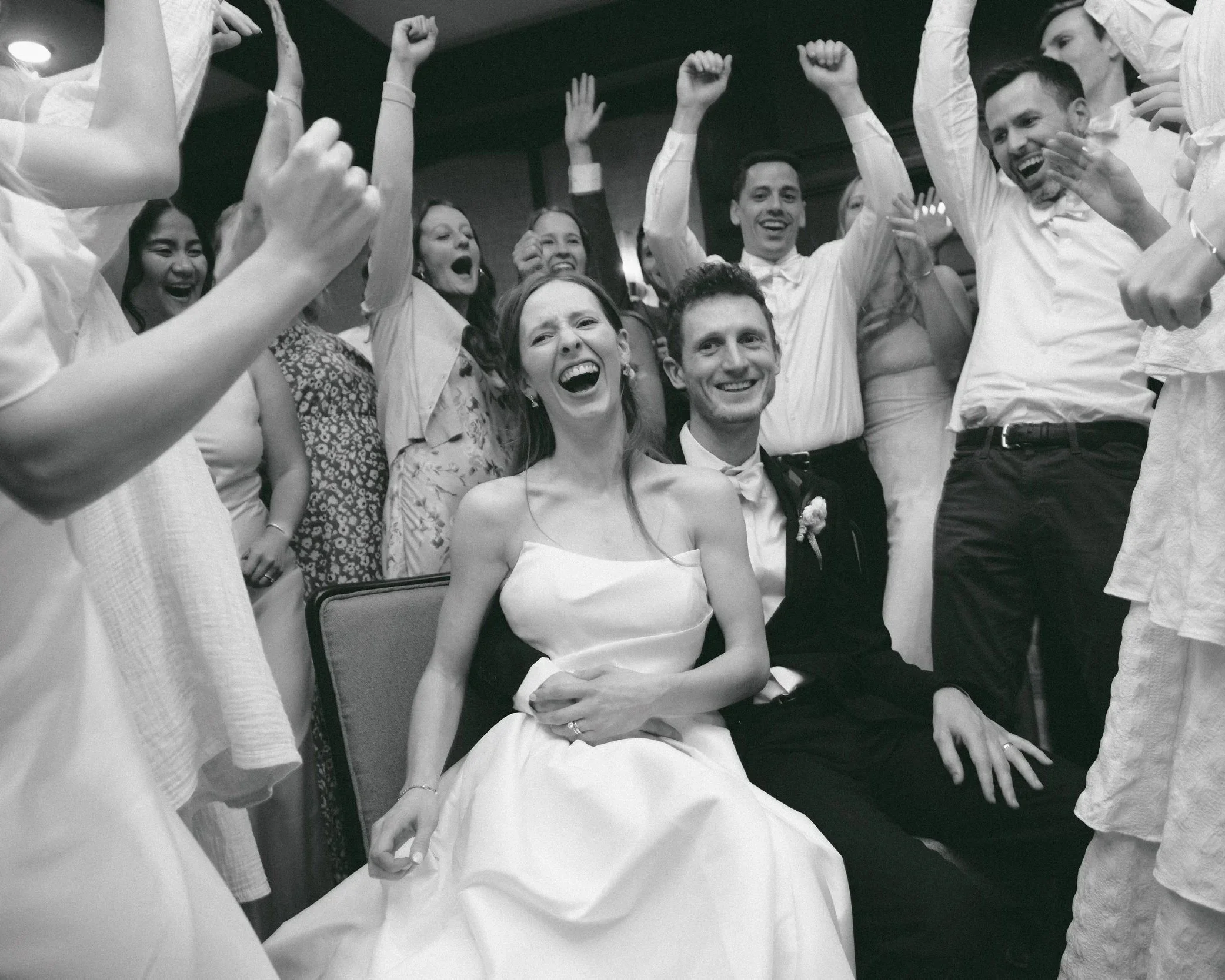 Black and white photo of a wedding celebration with a woman in a wedding dress and a man in a tuxedo seated in the center, surrounded by smiling guests with raised hands.