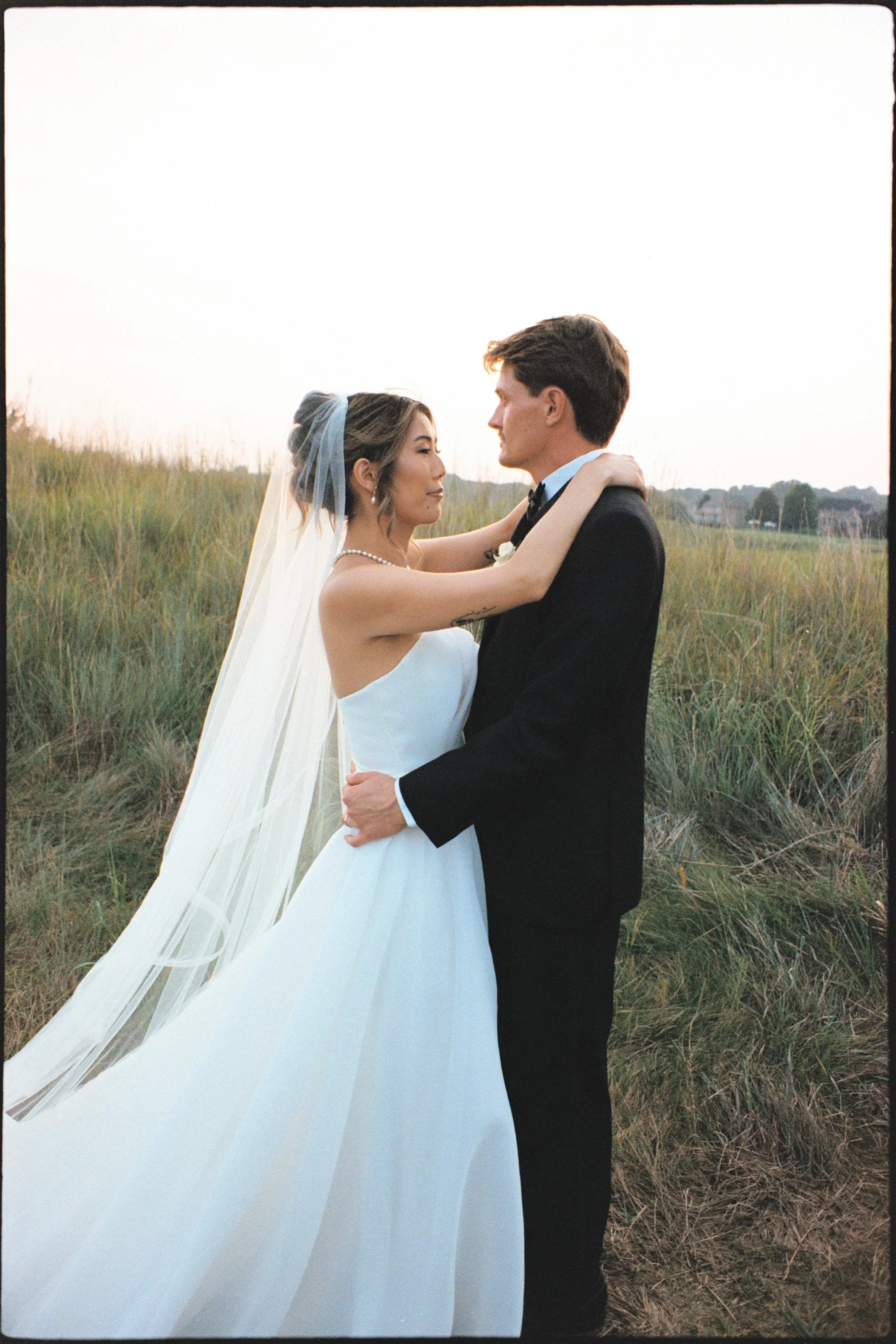 Bride and groom embracing outdoors during sunset, the bride in a strapless white wedding dress with a veil and pearl necklace, the groom in a black tuxedo, standing on grassy terrain.