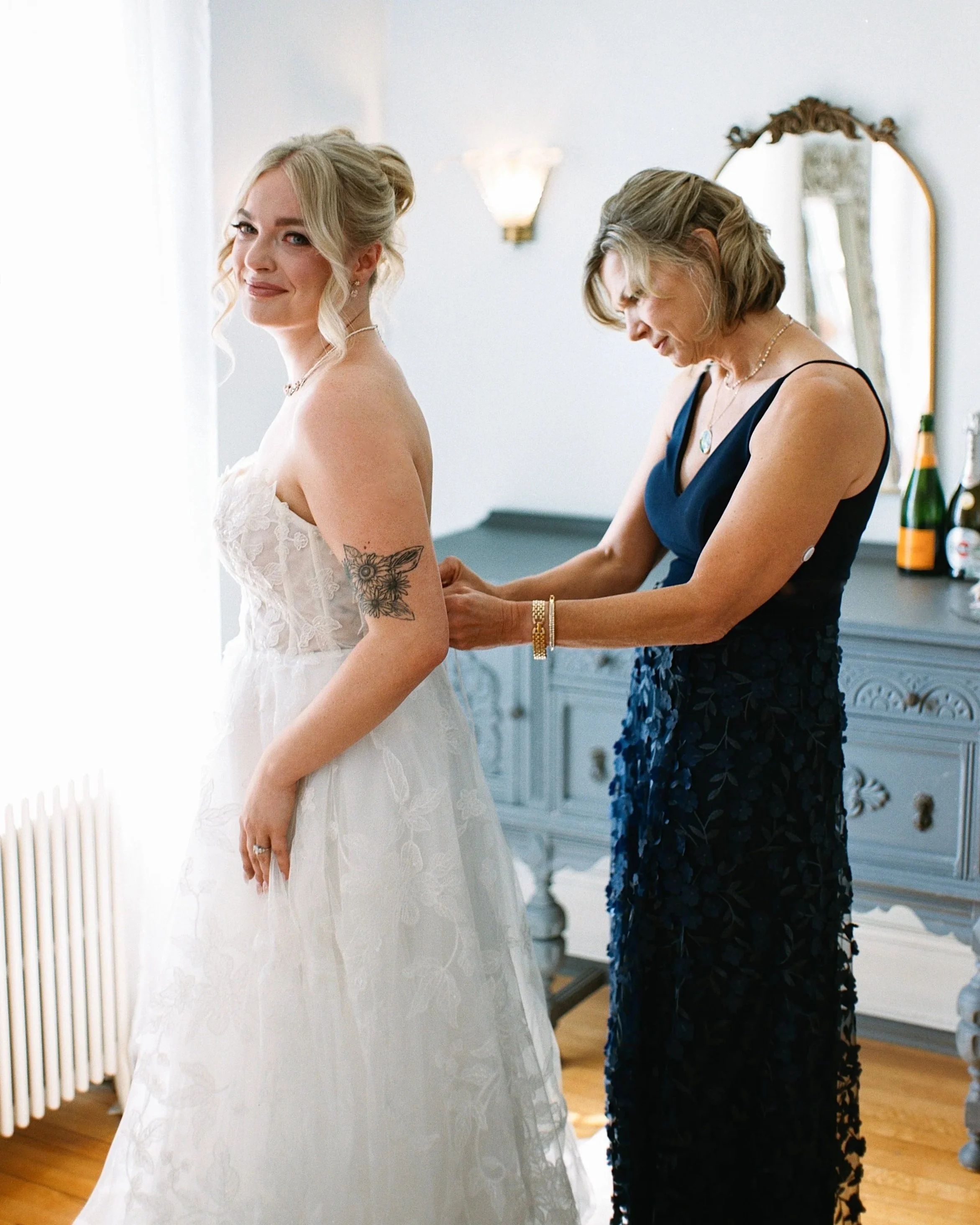 A bride in a white lace dress smiling as a woman helps her adjust her gown.