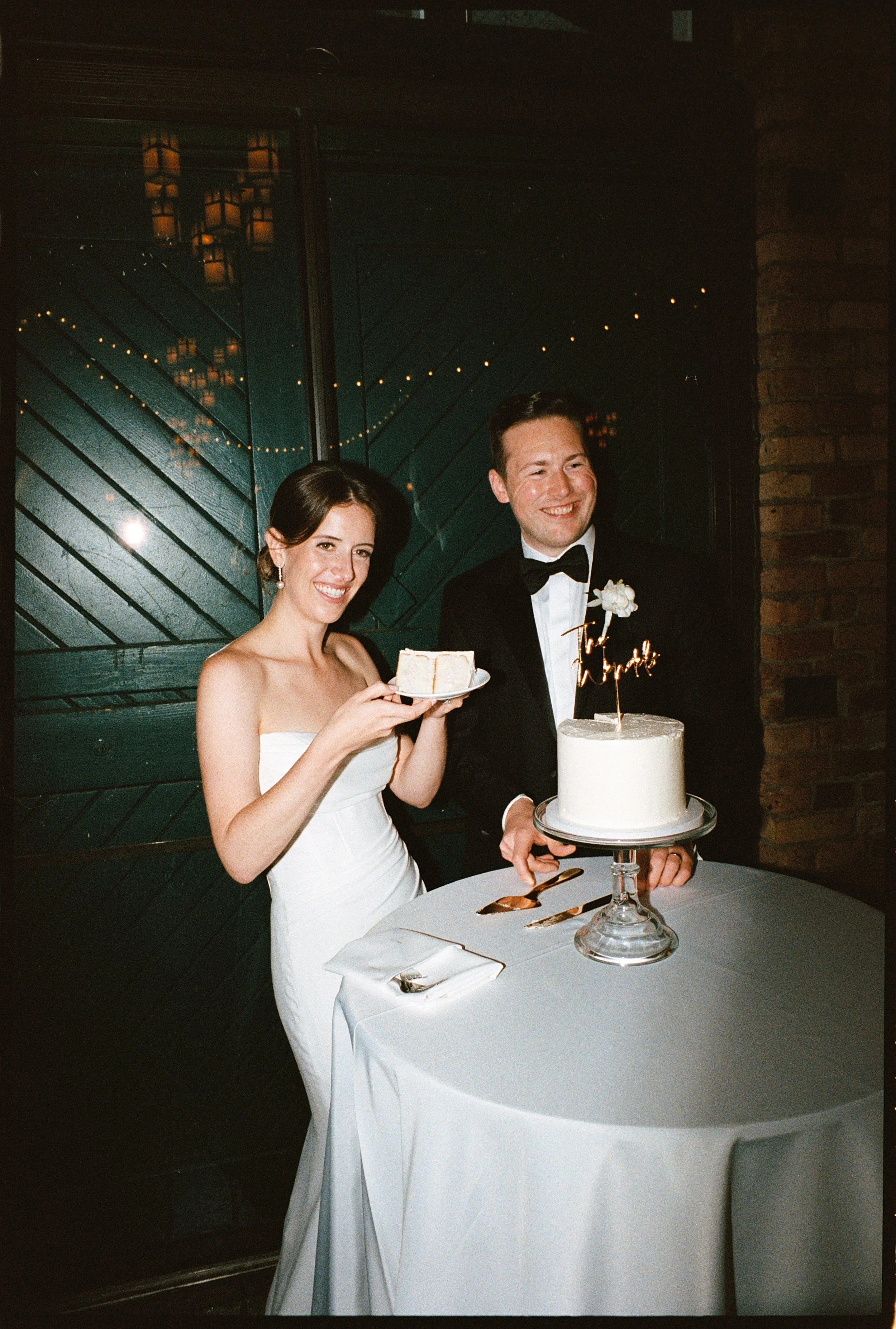 A newlywed couple at a wedding reception, smiling and standing next to a white wedding cake on a table. The bride is holding a plate with a slice of cake, and the groom is holding the cake with a 'Together' topper and a flower decoration.
