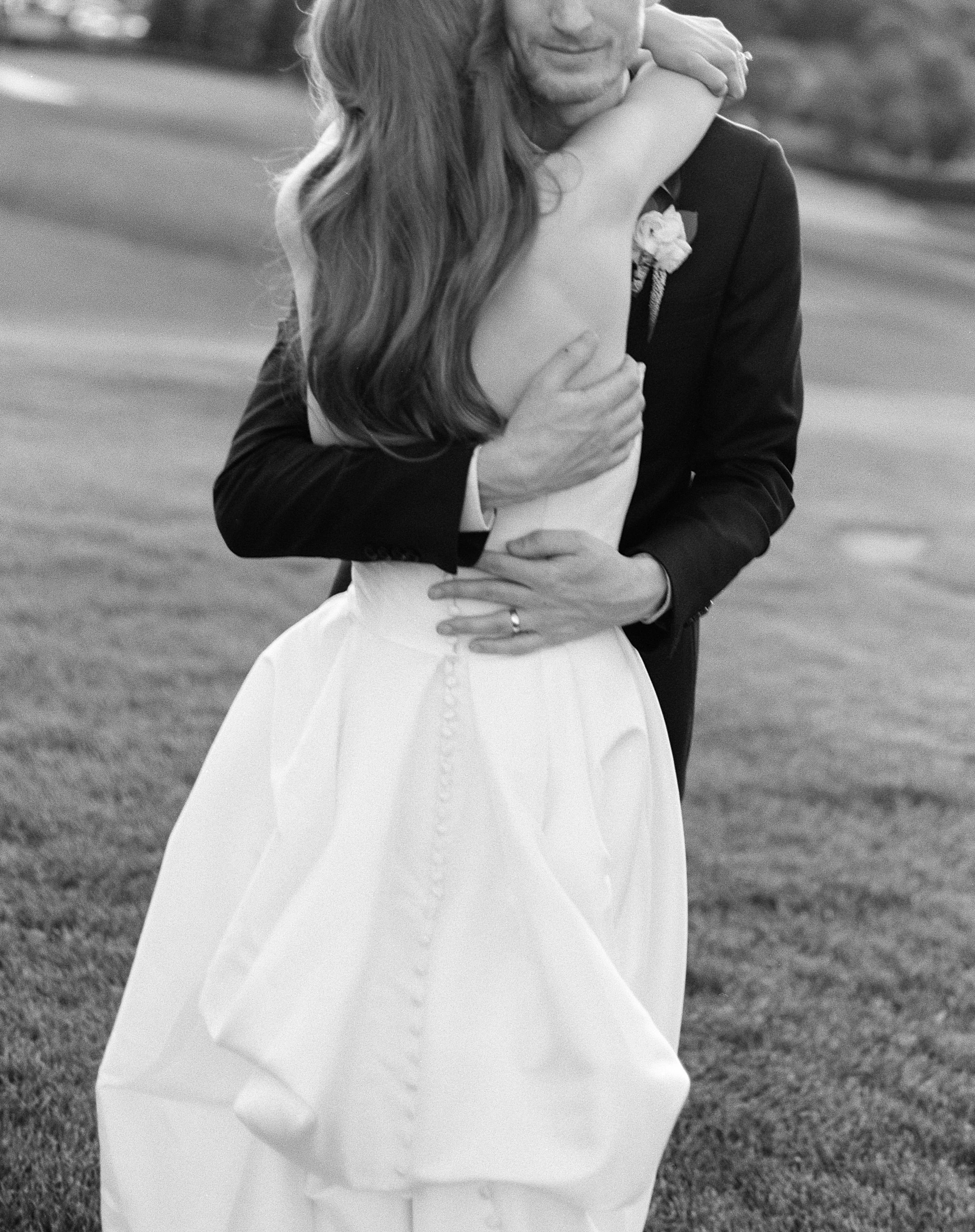 A black and white photo of a man and woman embracing each other outdoors, with the woman wearing a wedding dress and the man in a suit.