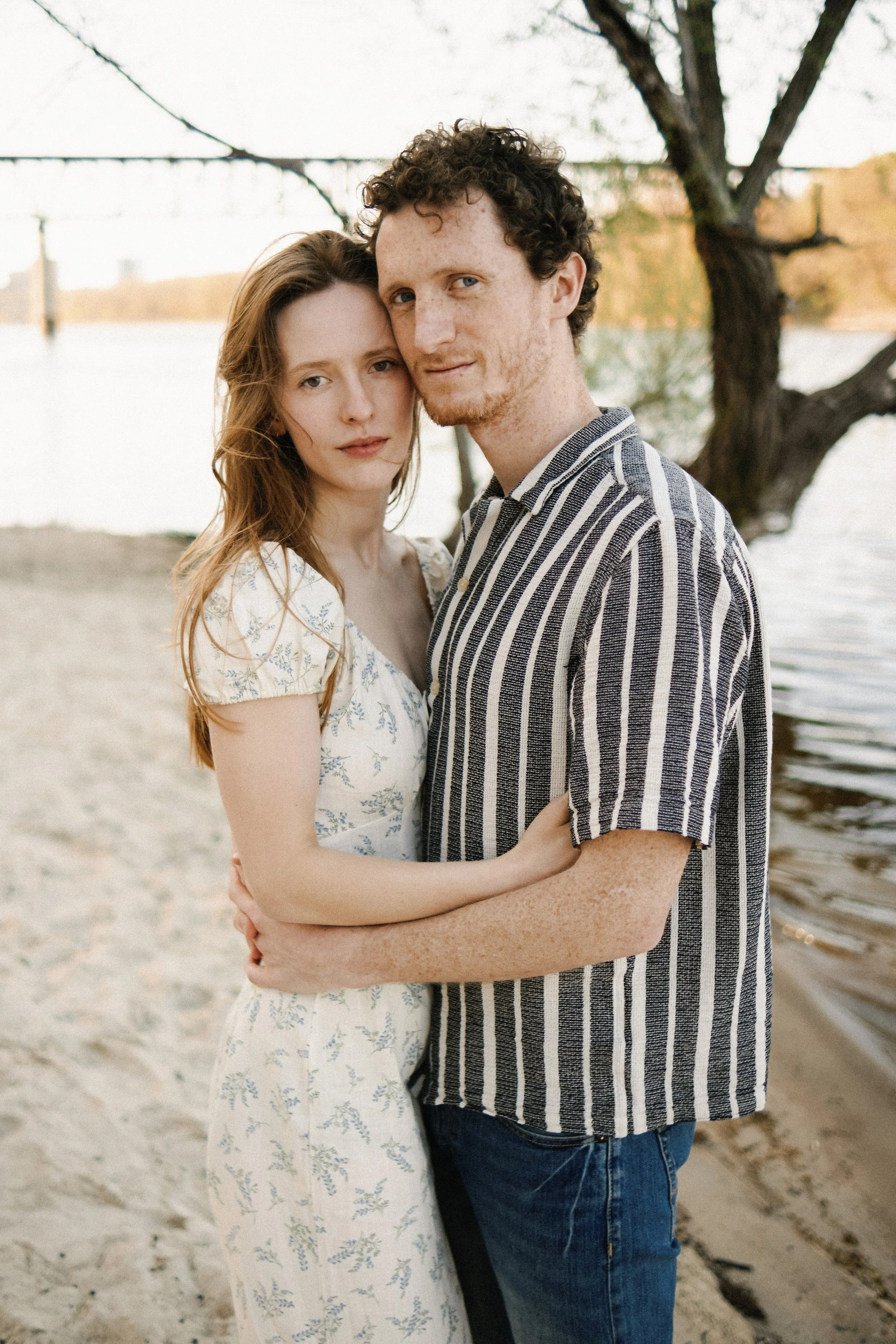 A young couple standing close together on a beach near water with trees in the background, embracing, during sunset.