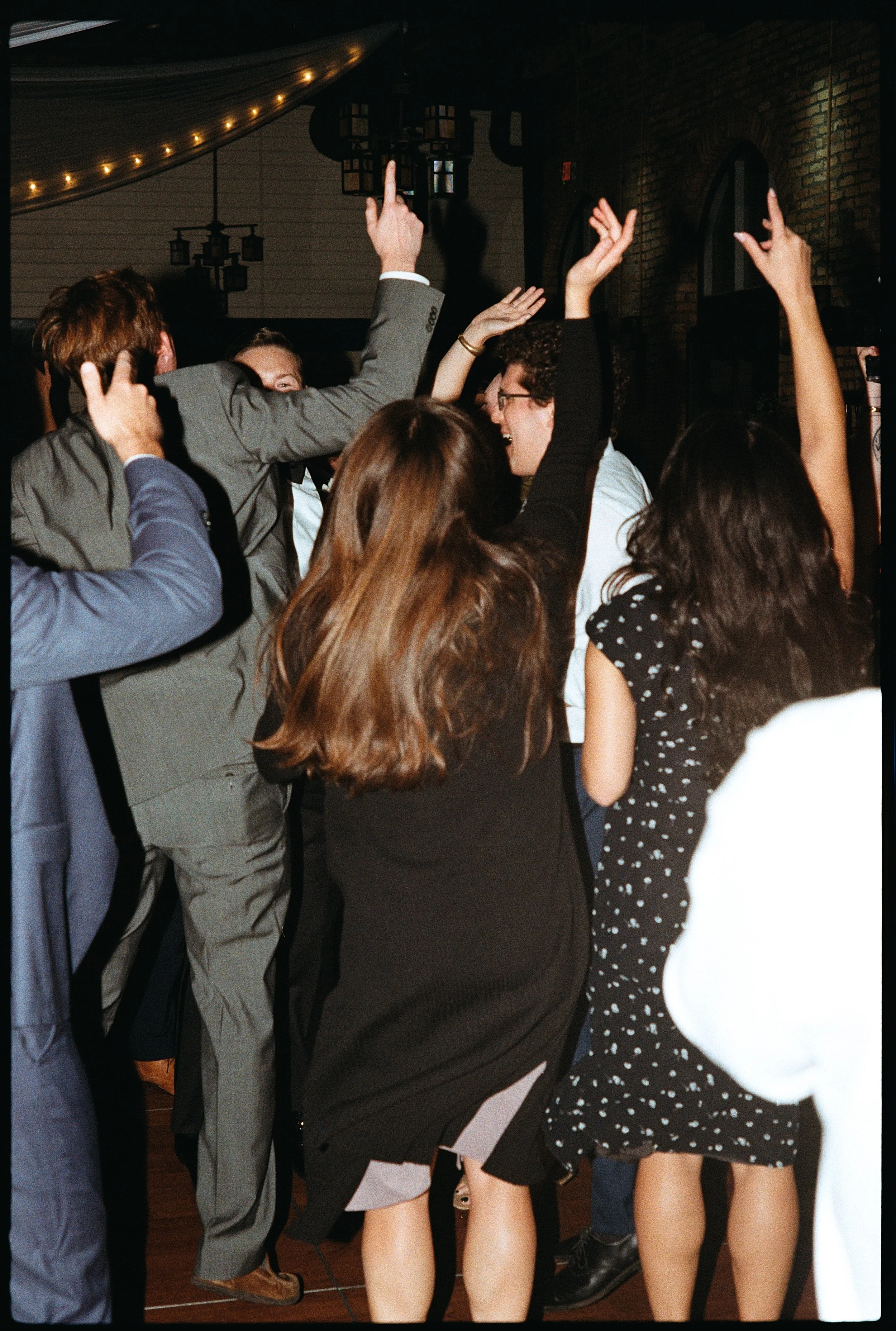 People dancing and celebrating at a party or event in a dimly lit venue with string lights and brick walls.
