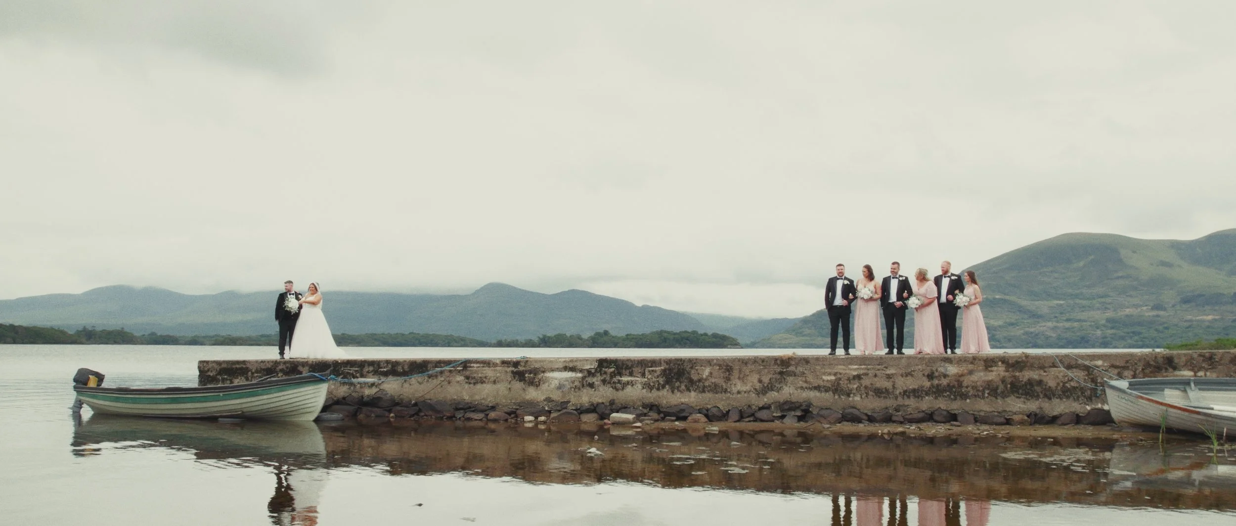 A wedding party on a stone pier by a lake, with mountains in the background. The bride and groom are on the left, and four bridesmaids and groomsmen are on the right, holding bouquets and dressed formally.