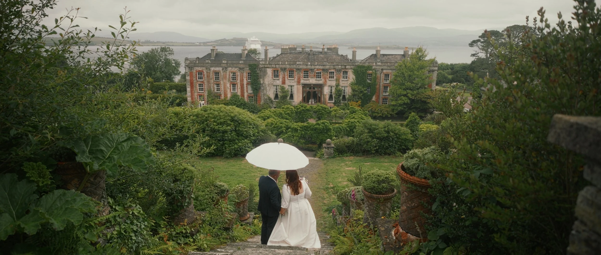 A bride and groom standing under a white umbrella on stone steps in a lush garden, with an elegant large historic mansion in the background during overcast weather.