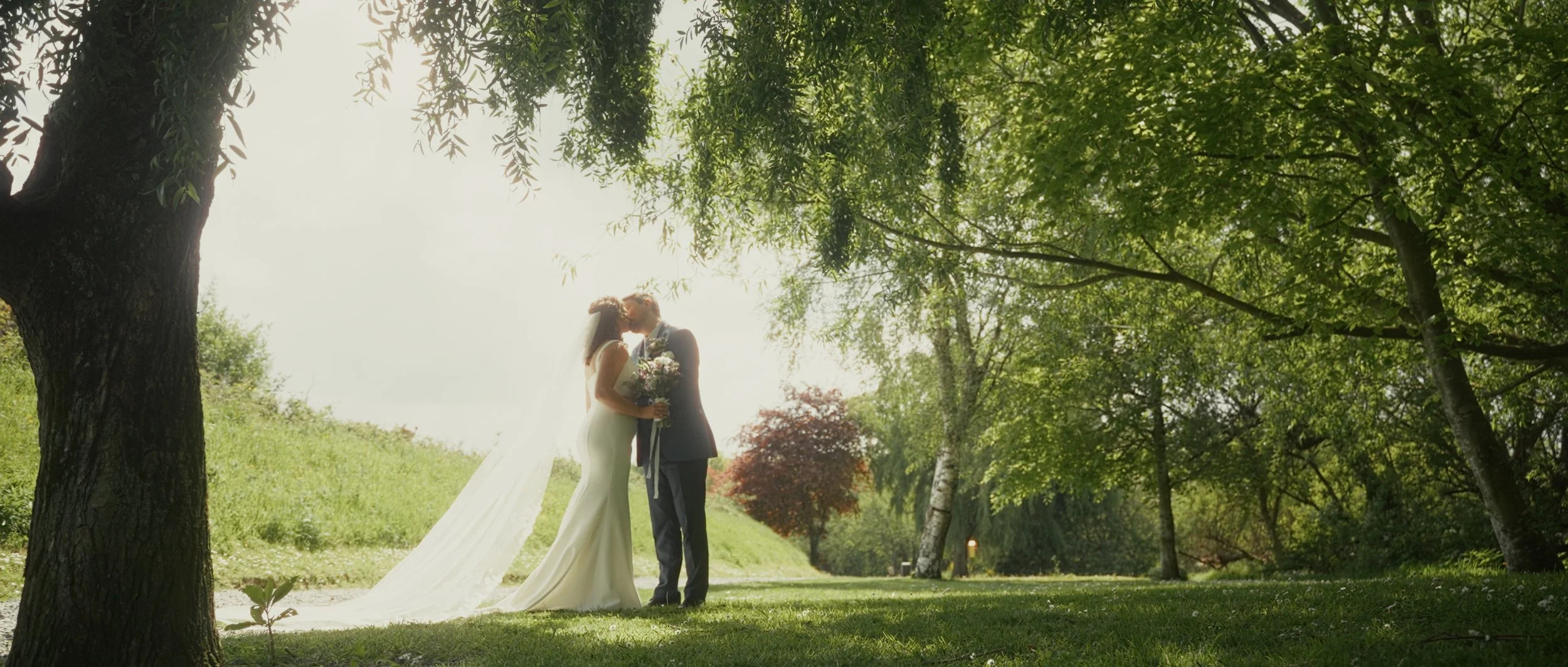 A bride and groom sharing a kiss under trees in a park during daytime, with sunlight filtering through the leaves.