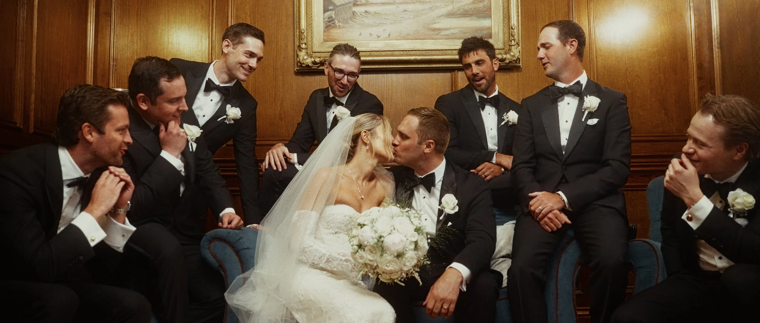 A bride and groom sharing a kiss surrounded by eight men in tuxedos at a wedding celebration.