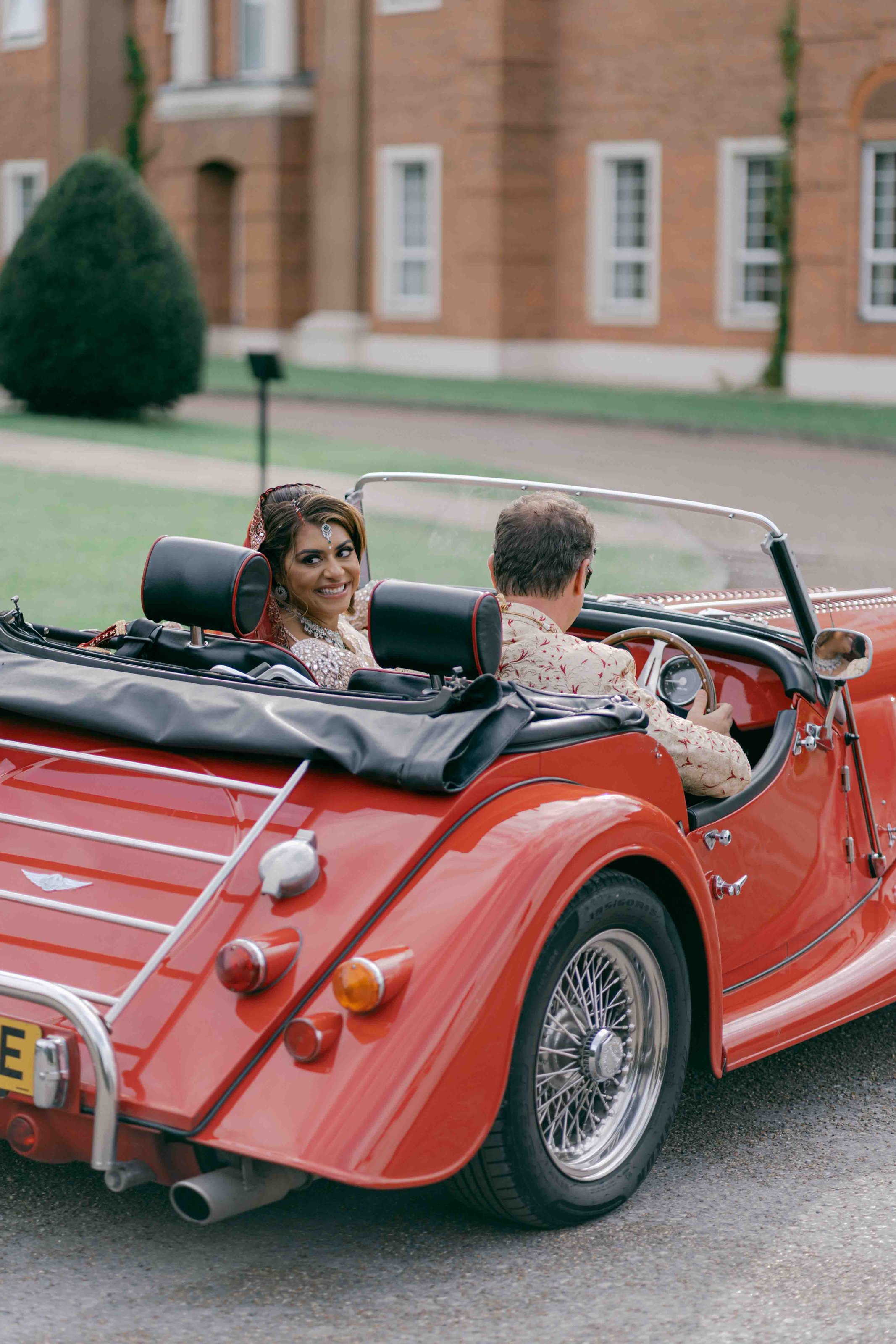 bride looking to the camera while groom is driving in a classic car 