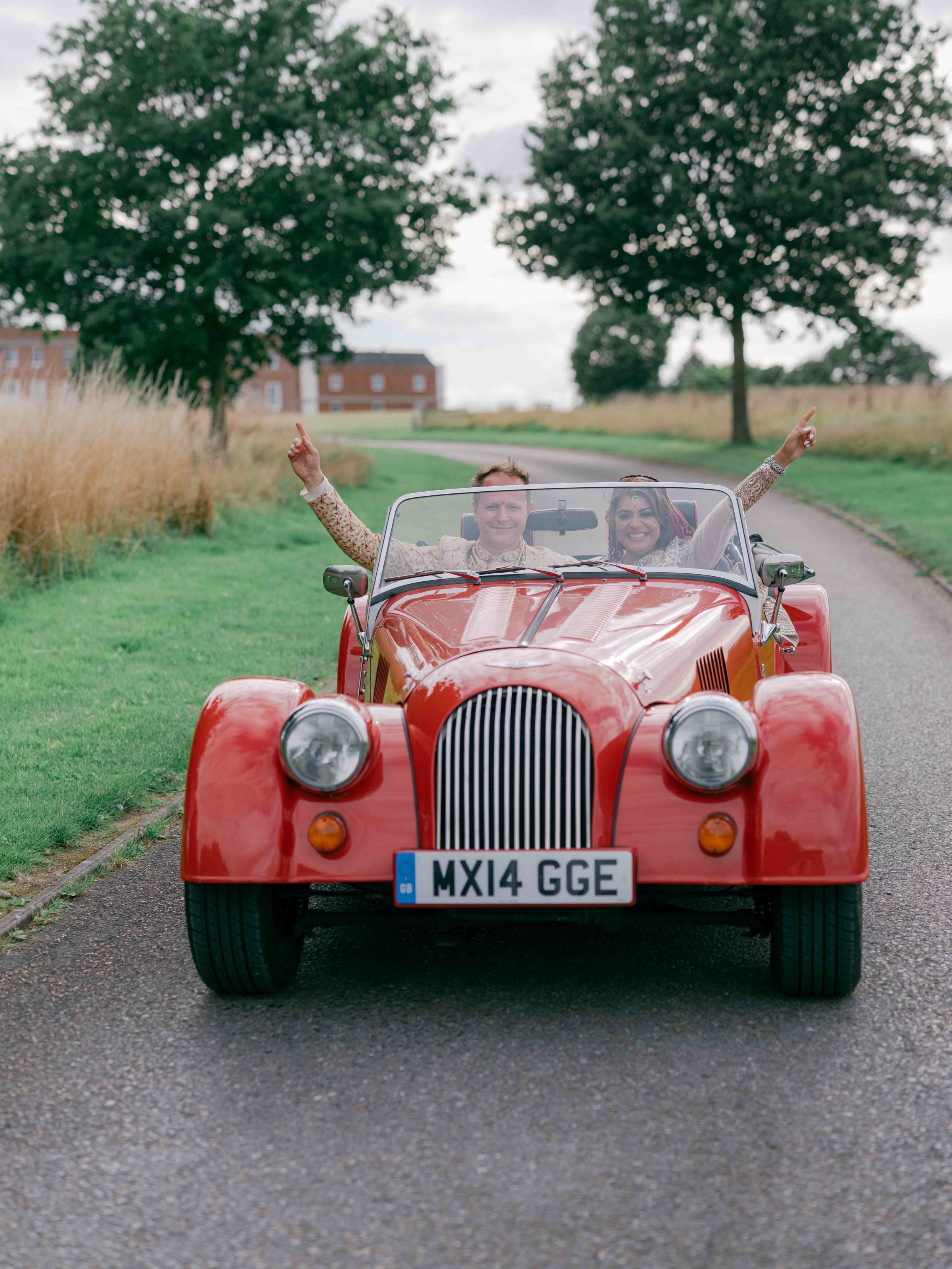  bride and groom driving toghther in a red classic car 