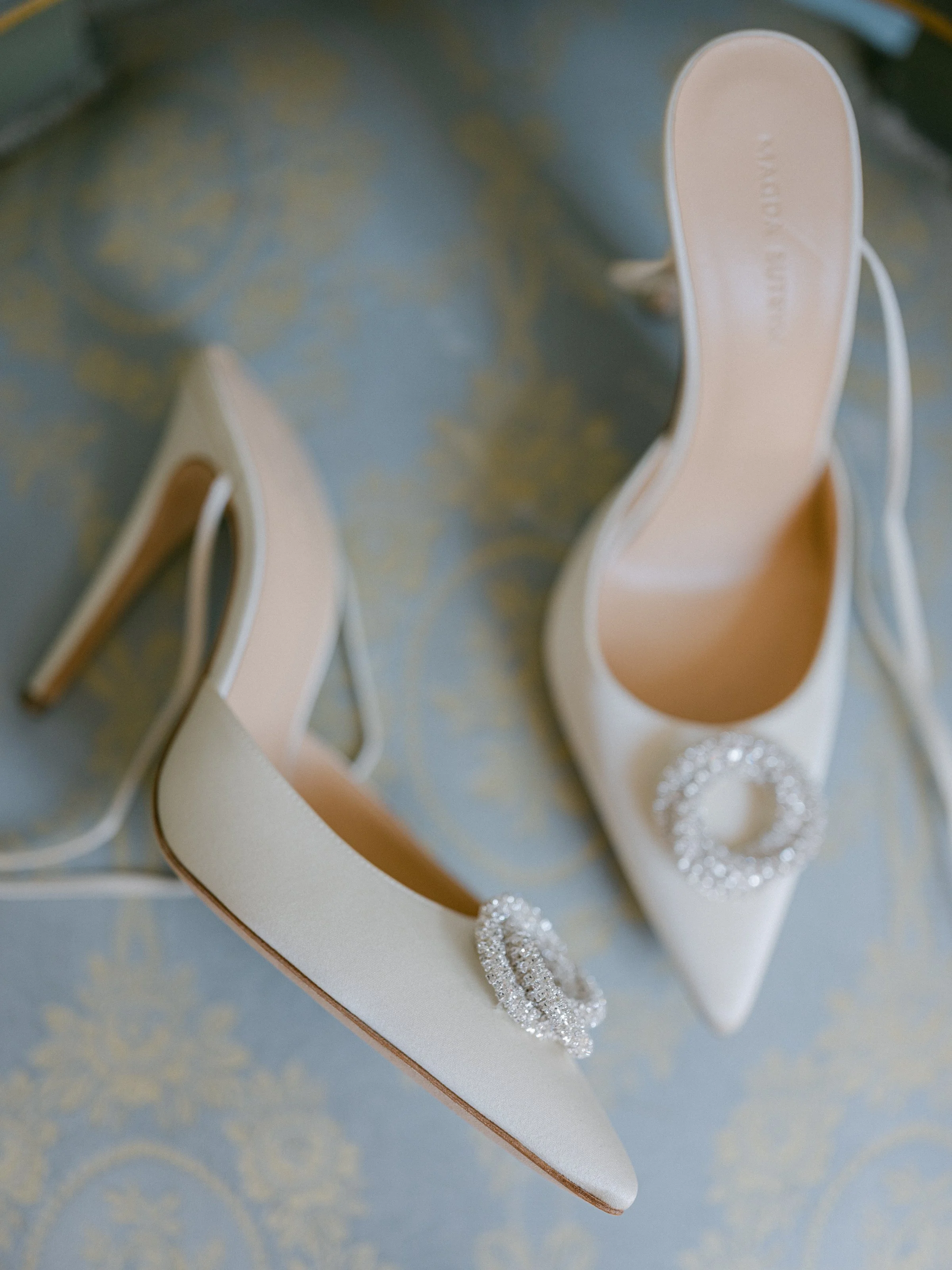 Pair of white high-heeled shoes with jeweled buckle decorations, placed on a decorative blue and gold patterned surface.