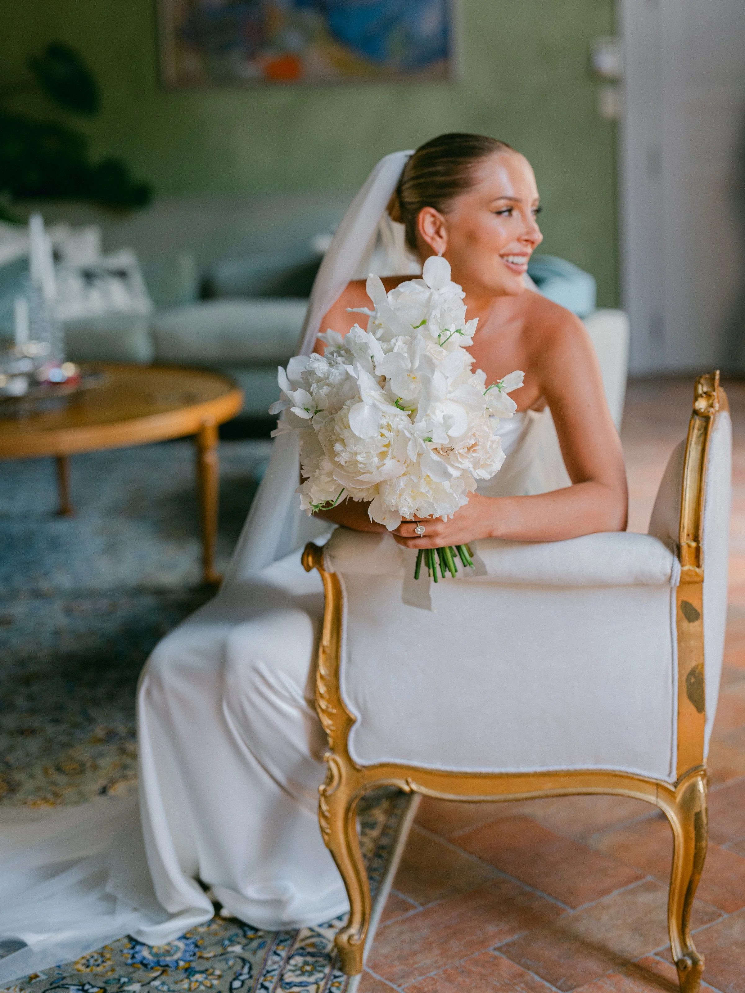 A bride in a white wedding dress sitting on a vintage armchair, holding a bouquet of white flowers, smiling and looking to her right in a warmly lit room.