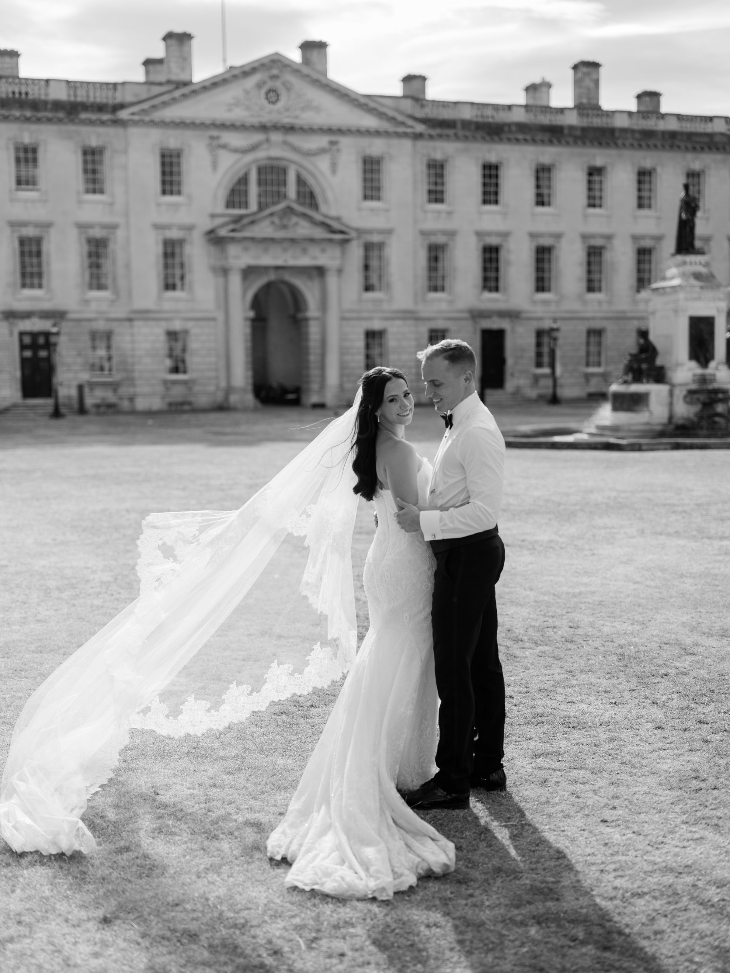 Black and white photo of a bride and groom embracing outdoors in front of a historic building, with the bride wearing a wedding gown and veil, and the groom in a tuxedo.
