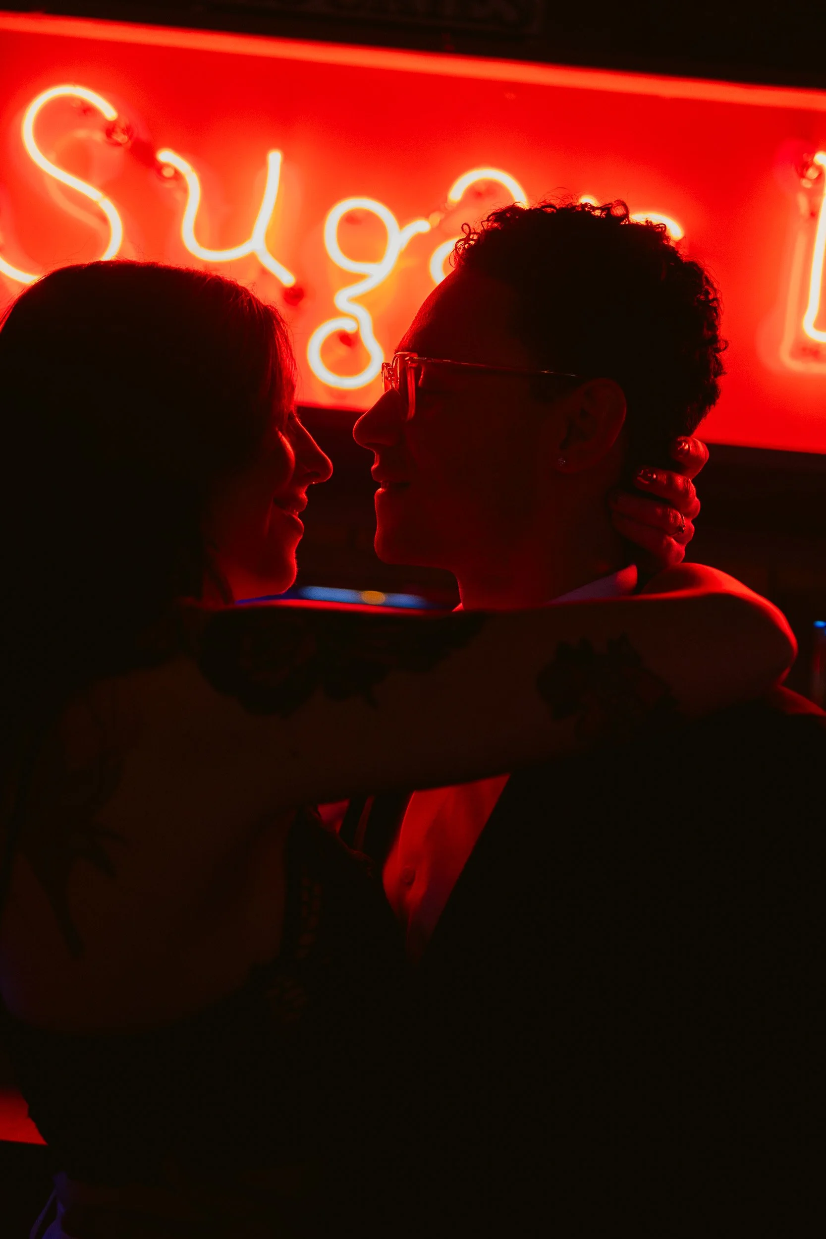 Man and woman in an embrace, with red lighting. She has her arms around his shoulders with her hand touching the back of his neck. They are engaged.
