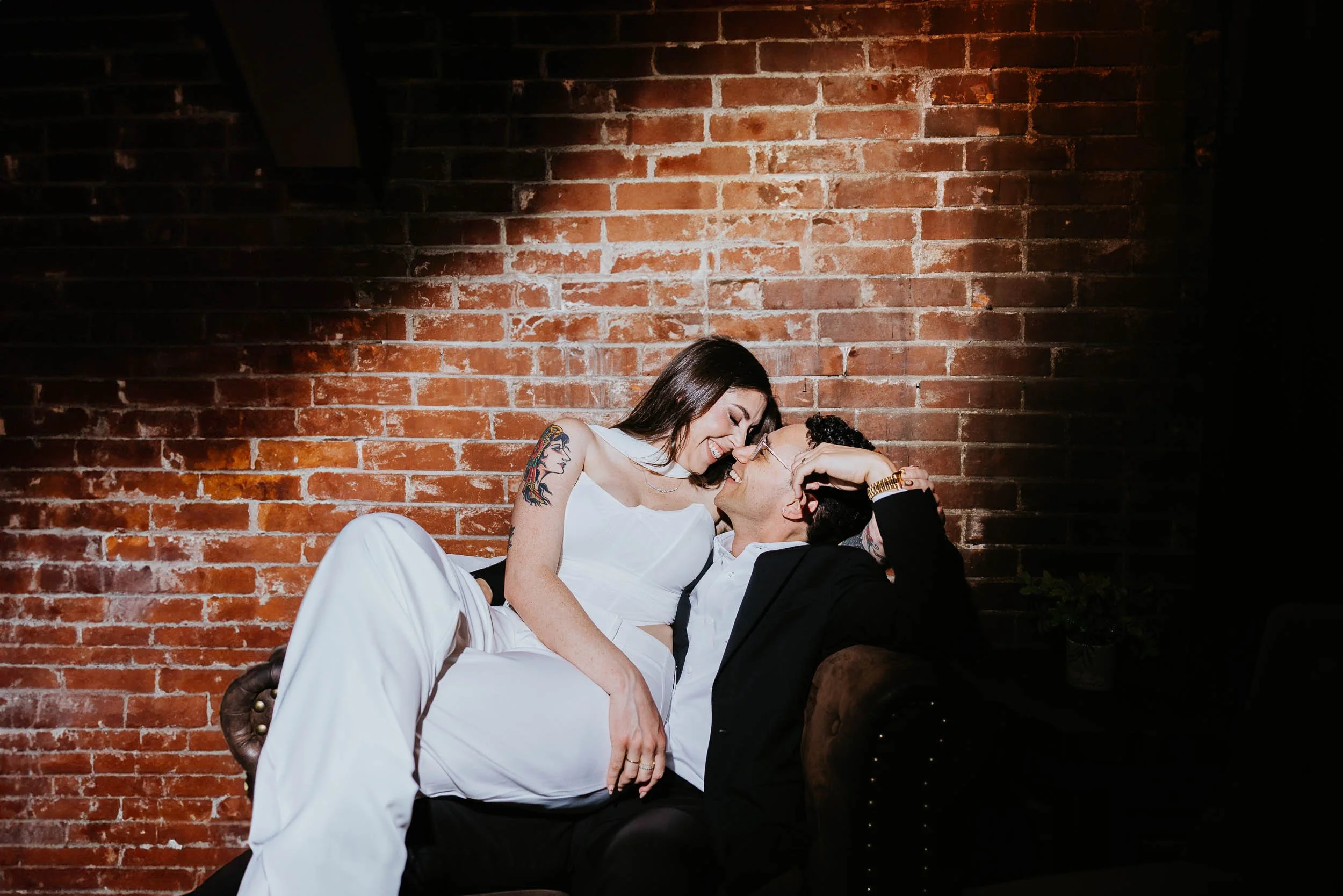 engaged couple sitting together on a chair, with a brick background and dramatic lighting. The woman is wearing white and sitting on his lap. He is wearing a black suit. They are smiling and almost kissing.