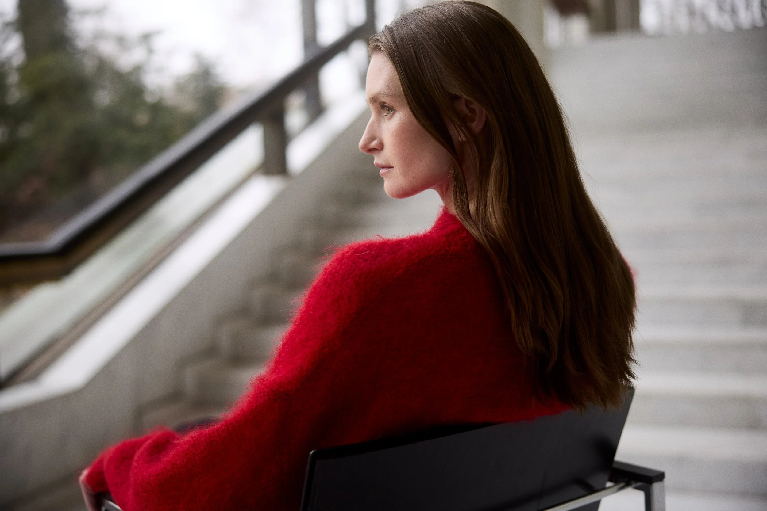 A woman with long brown hair, wearing a red sweater, sitting on a black chair on a staircase, looking to the left.