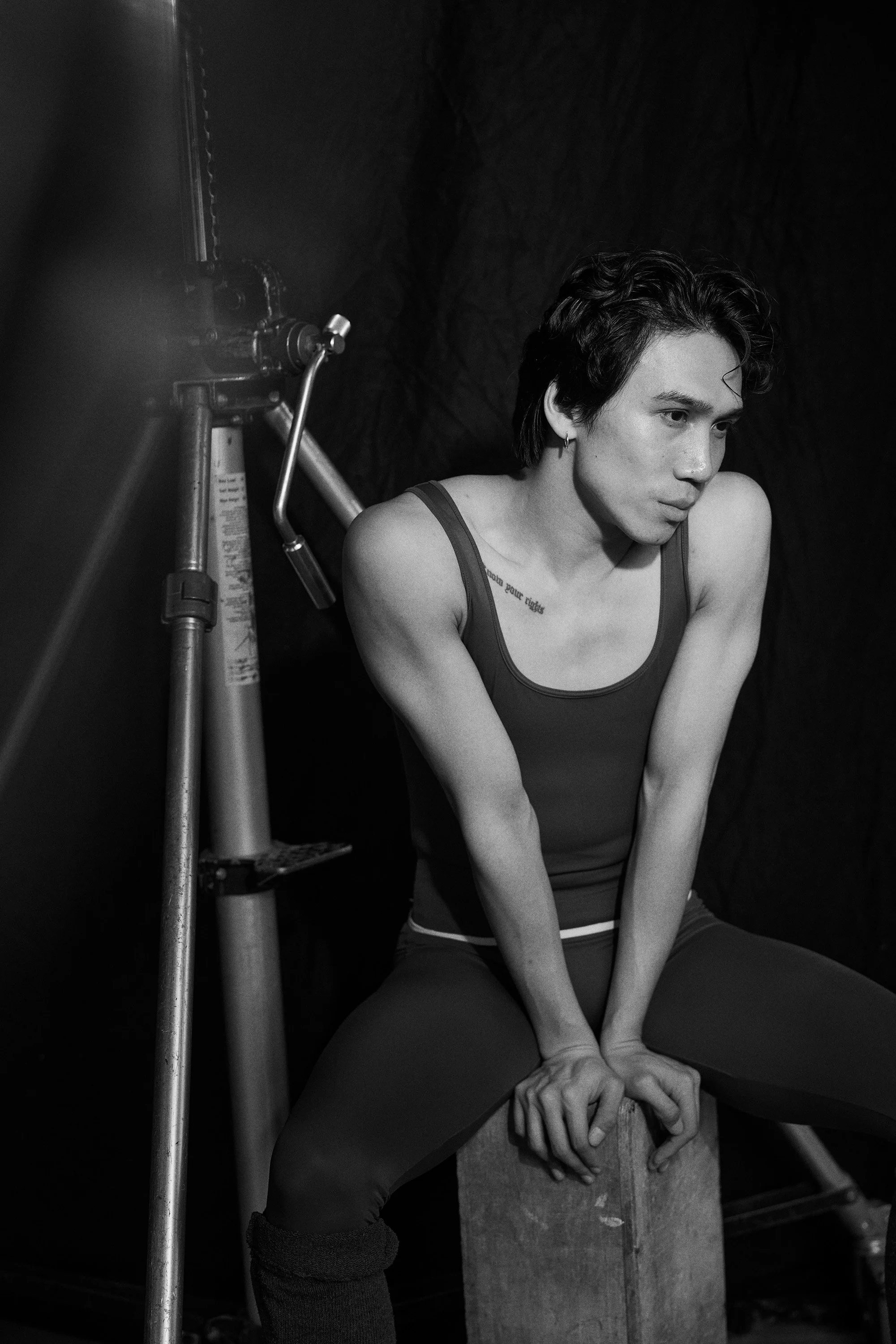 Black and white photo of a young woman with short dark hair, sitting on a wooden block, leaning forward with her hands clasped between her legs. She wears a tank top and dark pants, with a tattoo on her chest. There is photography equipment next to h