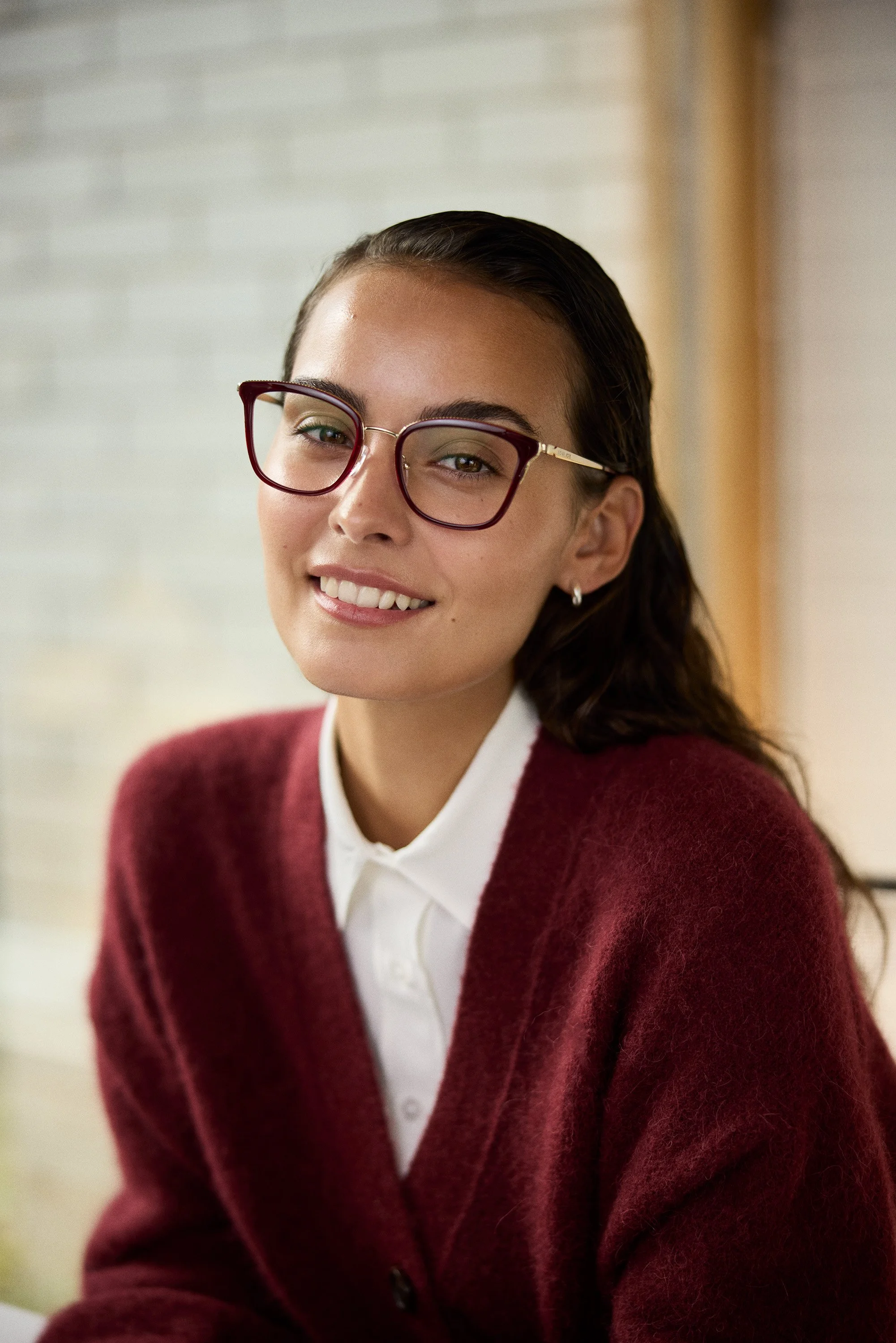 A young woman with brown hair, wearing glasses, a white shirt, and a maroon sweater, smiling at the camera in an indoor setting.