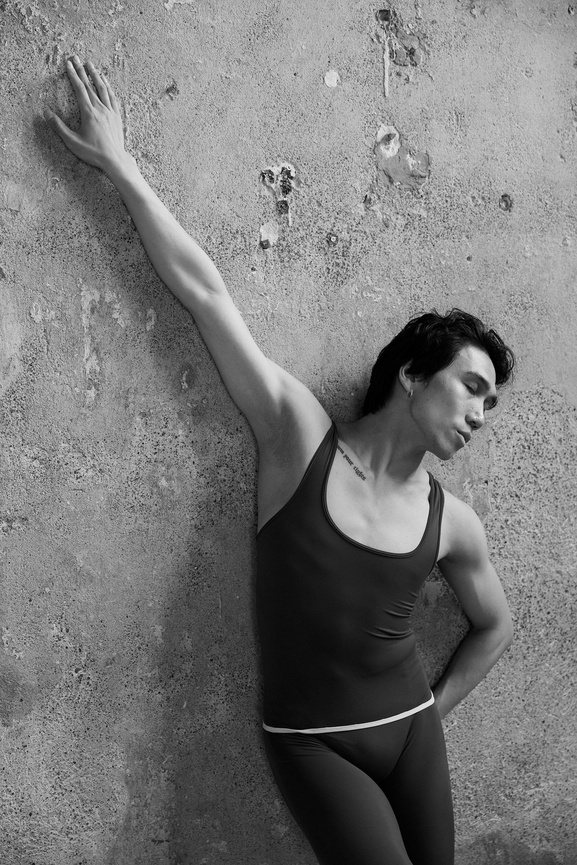 A woman in a sports tank top and leggings leaning against a textured wall with her eyes closed, one arm stretched out and touching the wall.